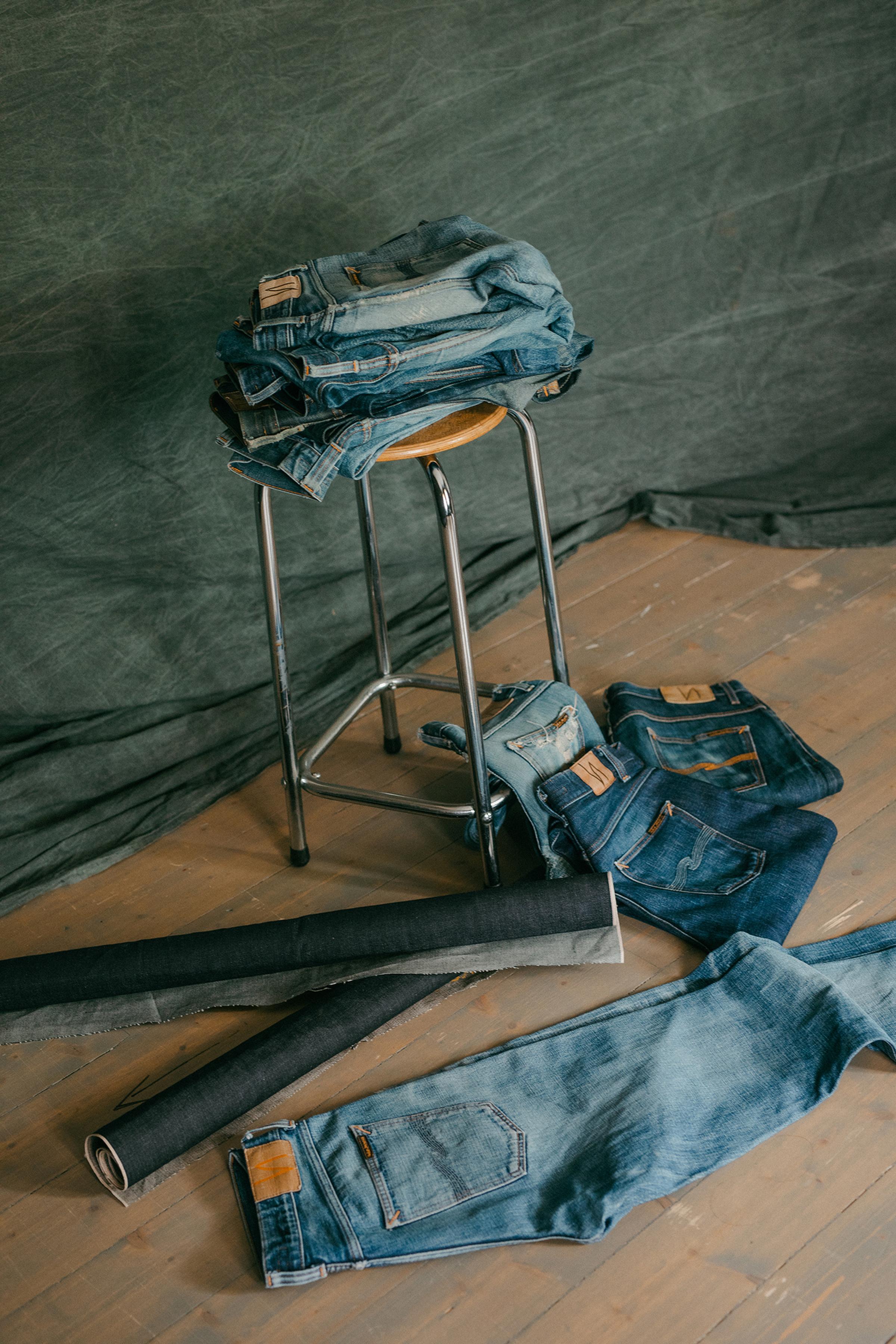 A stack of folded jeans on a stool, with more jeans and fabric rolls scattered on a wooden floor against a dark backdrop.