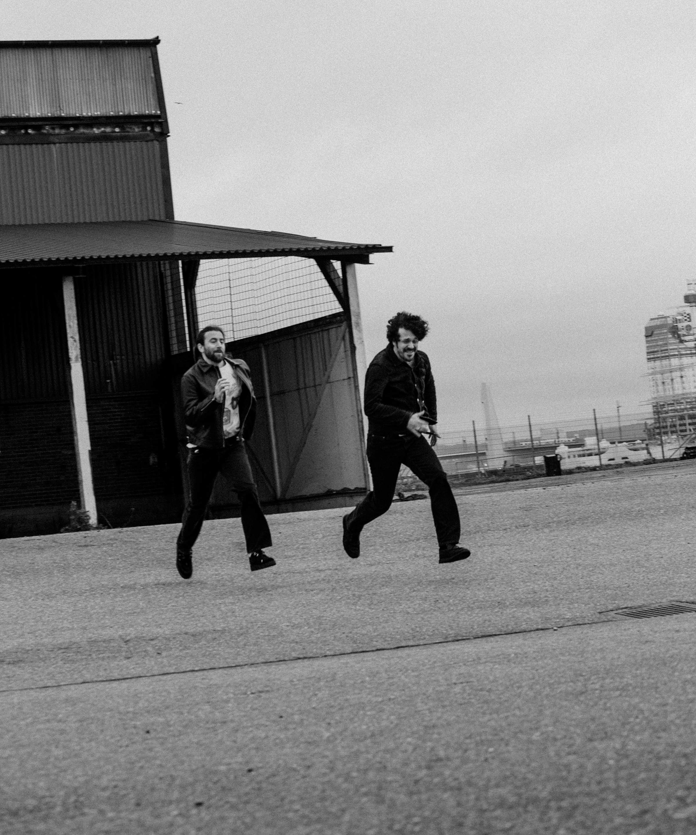 Two people running joyfully in an industrial area with a corrugated metal building and overcast sky in the background. Black and white photo.