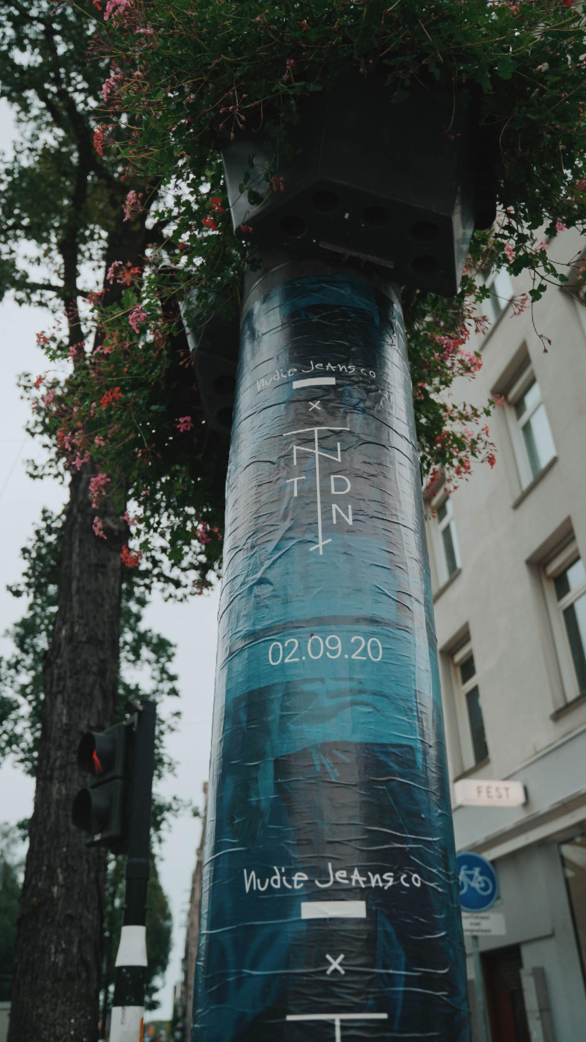A poster on a pole with a blue abstract design and text, surrounded by trees and buildings.