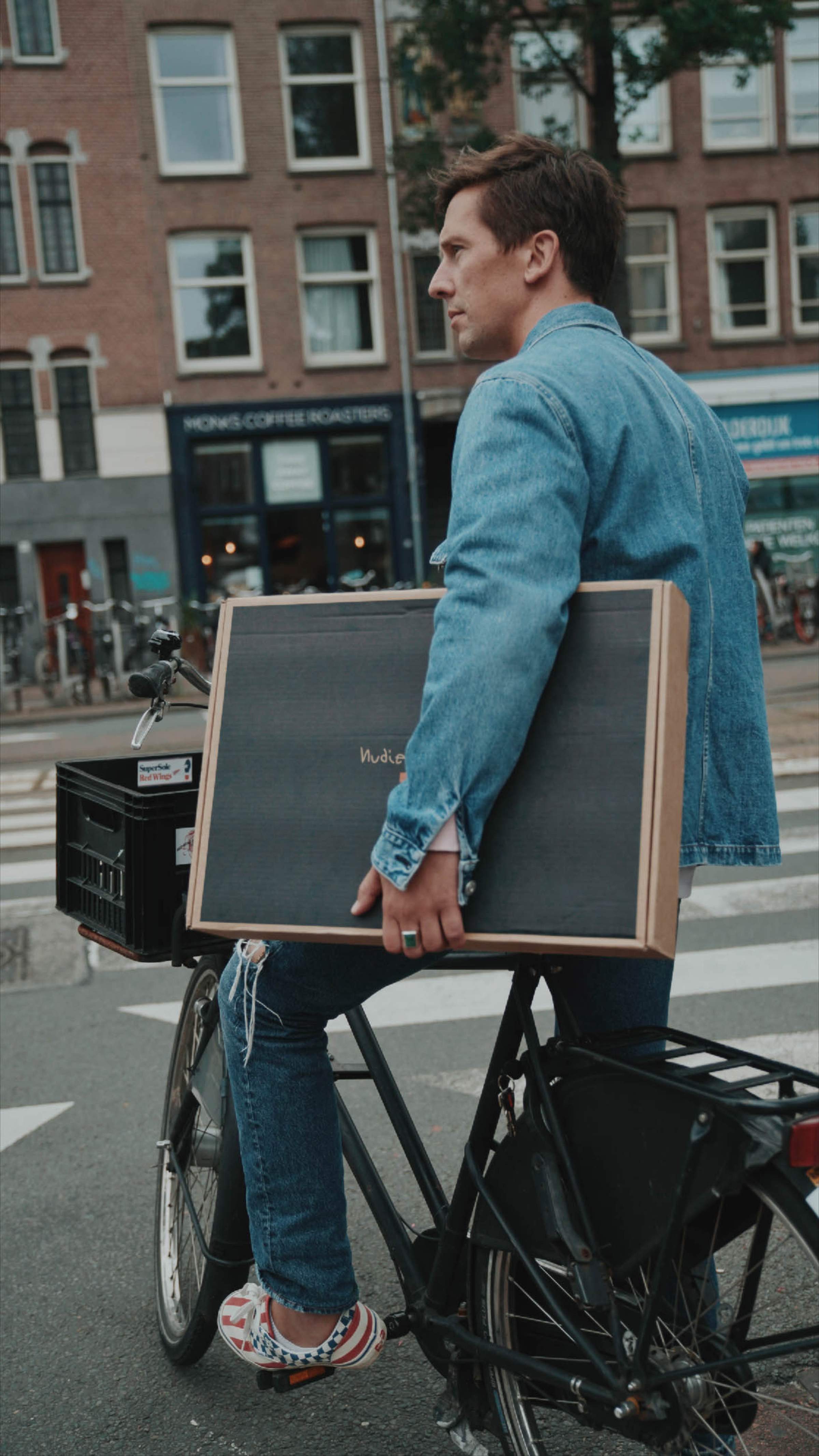 Man in denim jacket and jeans carries a nudie jeans box while riding a bicycle on a city street.