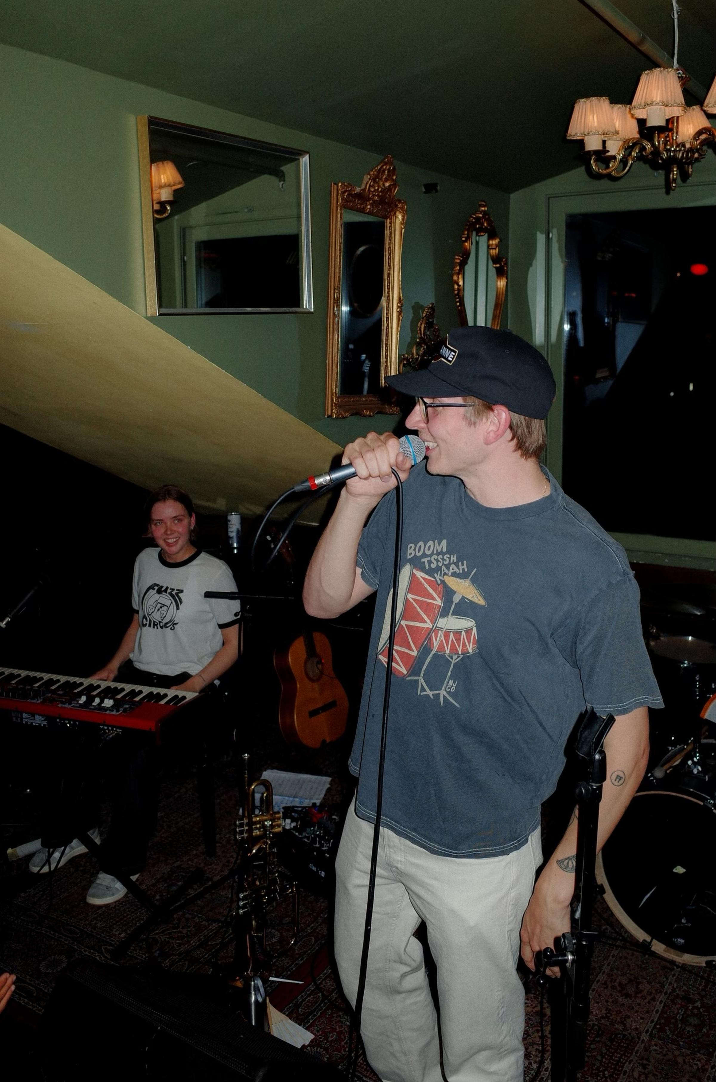 Mattis performs on a dimly lit stage with a musicians, and instruments. The audience's hands are visible in the foreground.