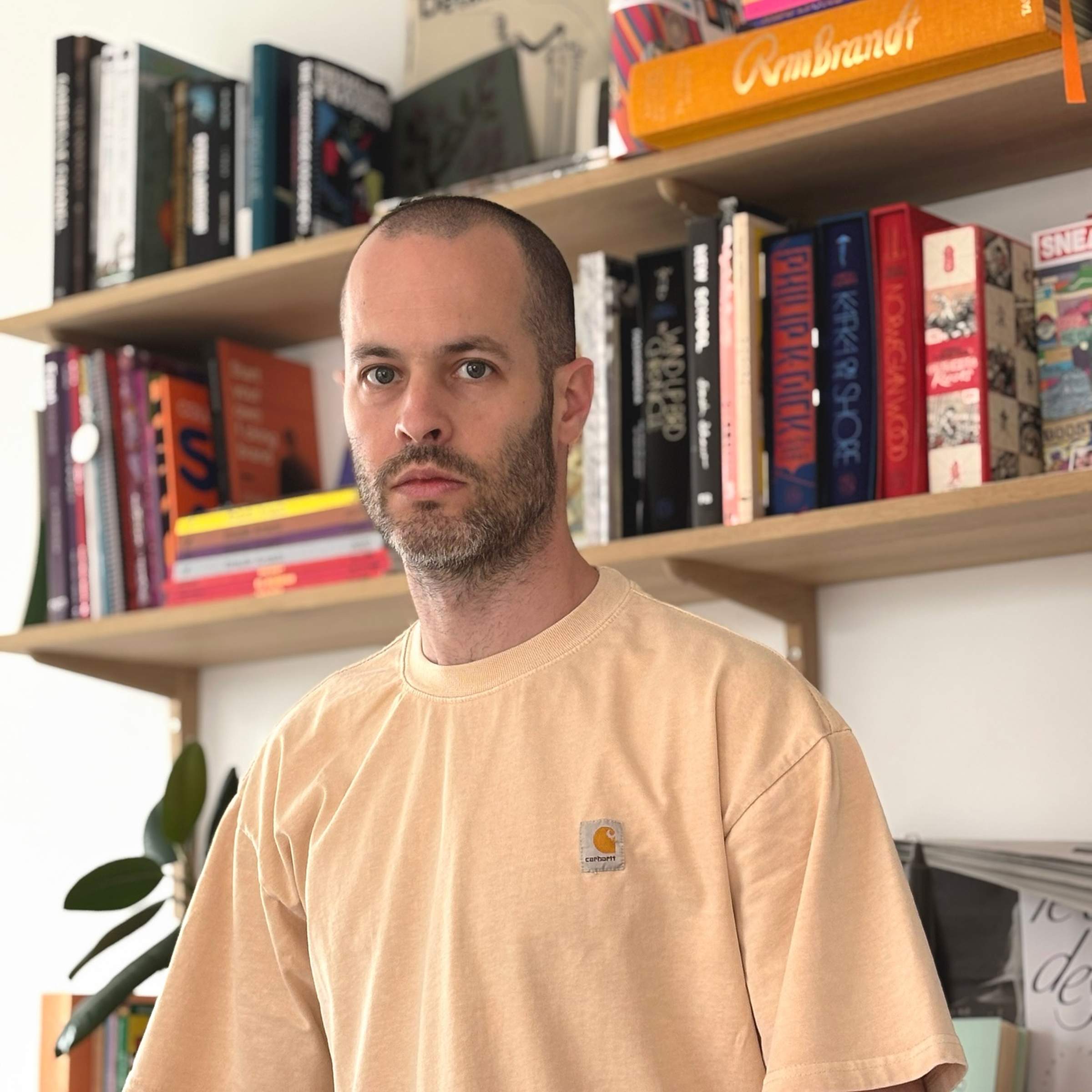 A person with wearing a beige T-shirt stands in front of bookshelves filled with colorful books.