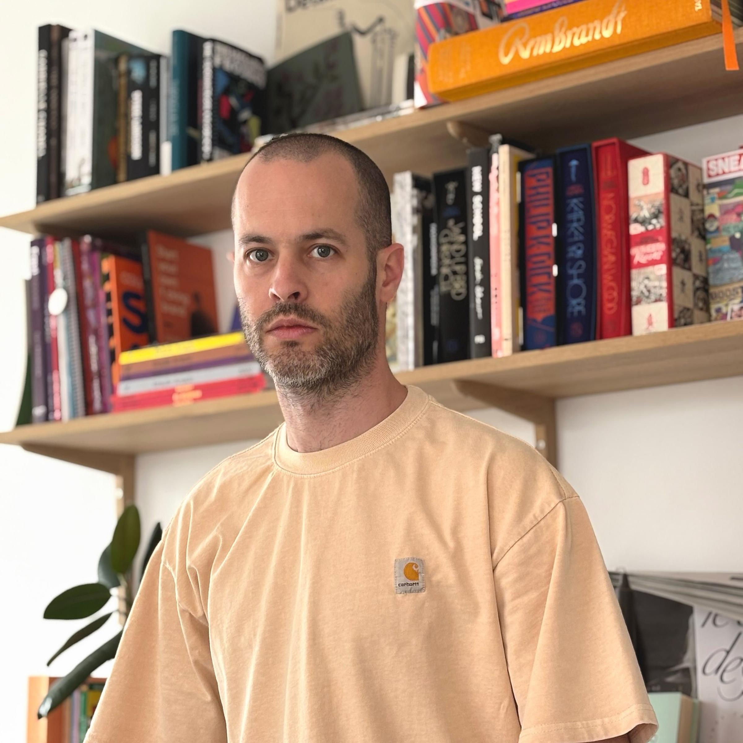 A person with wearing a beige T-shirt stands in front of bookshelves filled with colorful books.