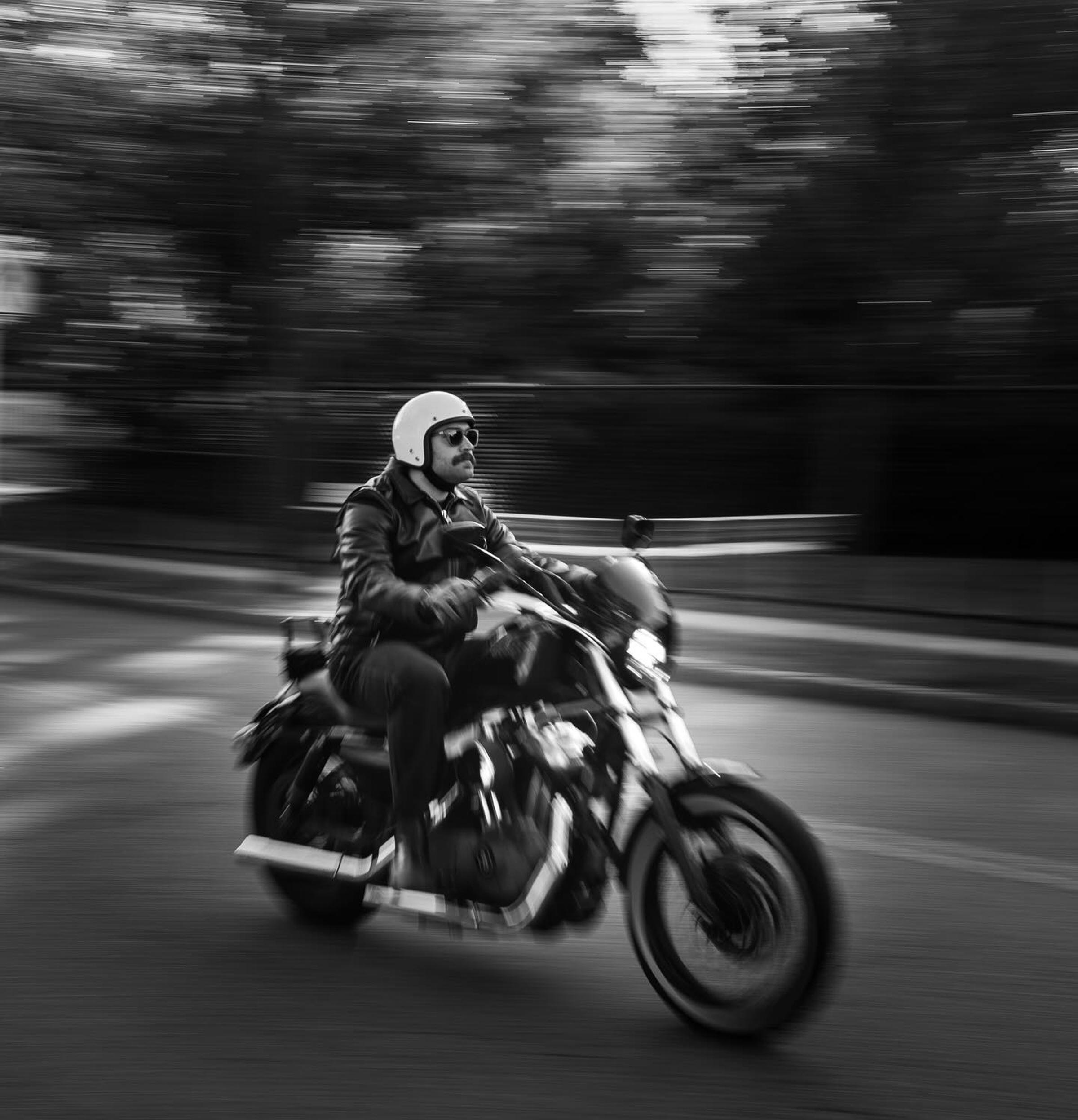 Black and white image of a person in a helmet riding a motorcycle at speed, with a blurred background indicating motion.