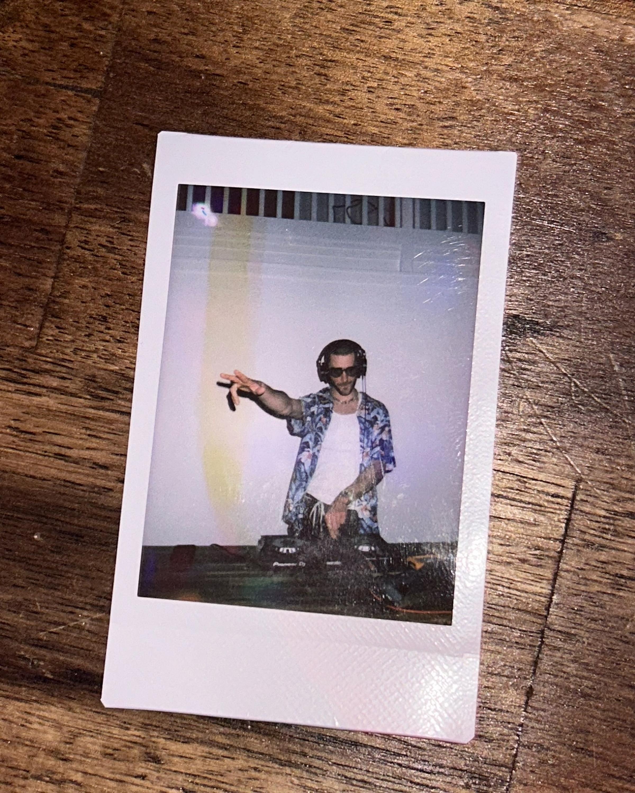 Polaroid of a DJ wearing sunglasses and a floral shirt, standing at a mixing console, posing with a peace sign.