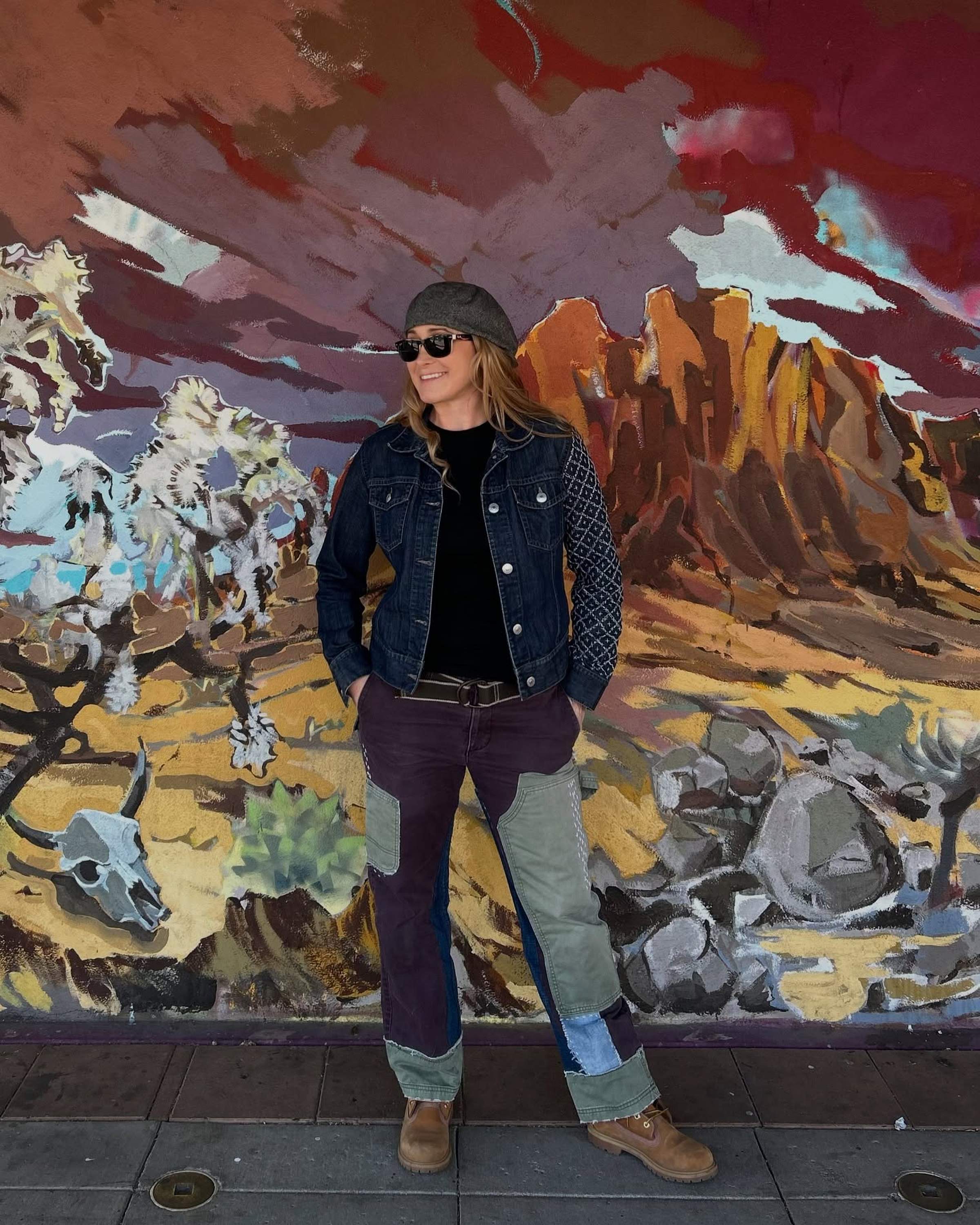 Woman in denim jacket and patchwork pants stands confidently before a colorful mural of a desert landscape, wearing sunglasses and a cap.