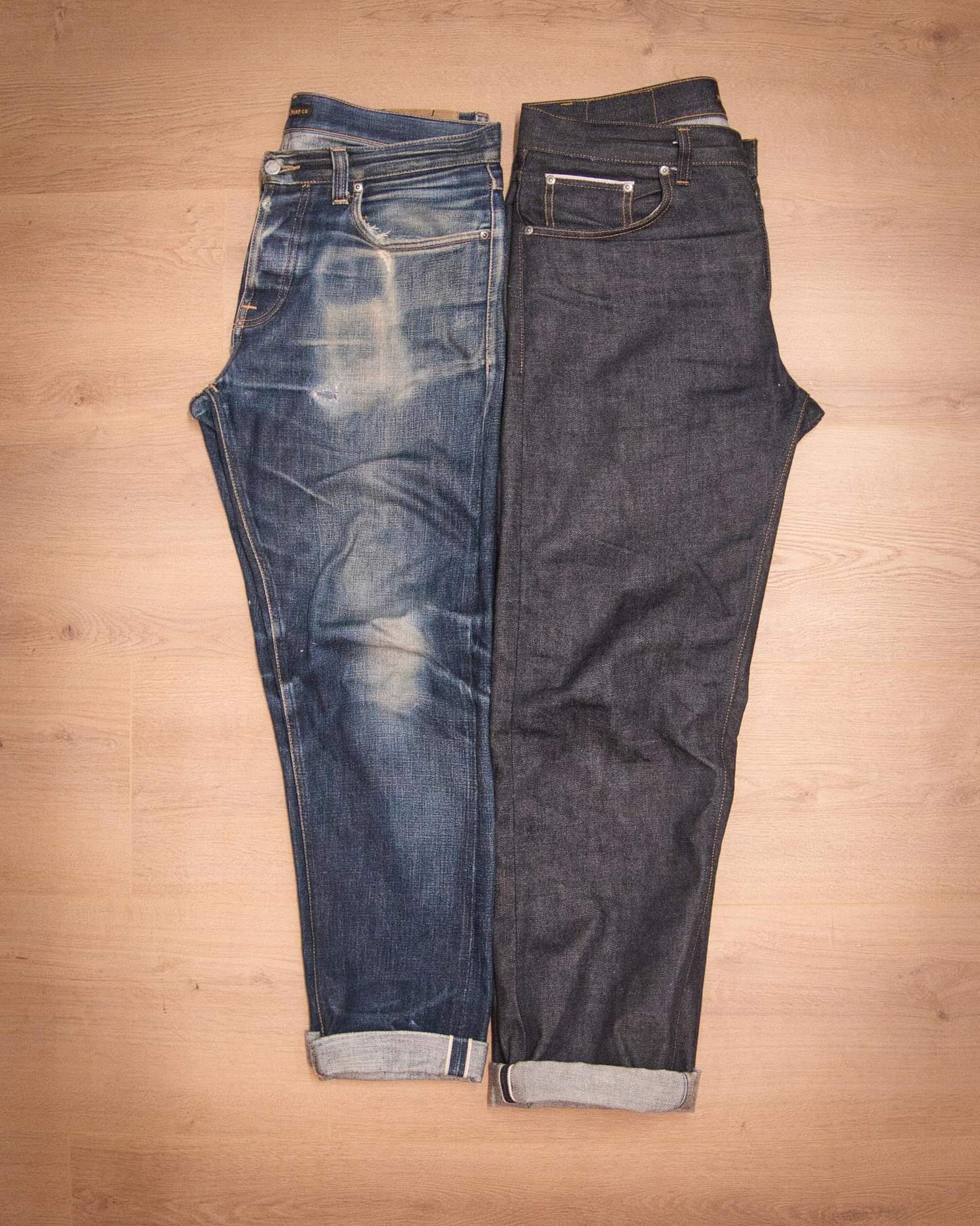Two pairs of Nudie jeans lie side by side on a wooden floor, one faded and distressed, the other dark and new, both with rolled cuffs.
