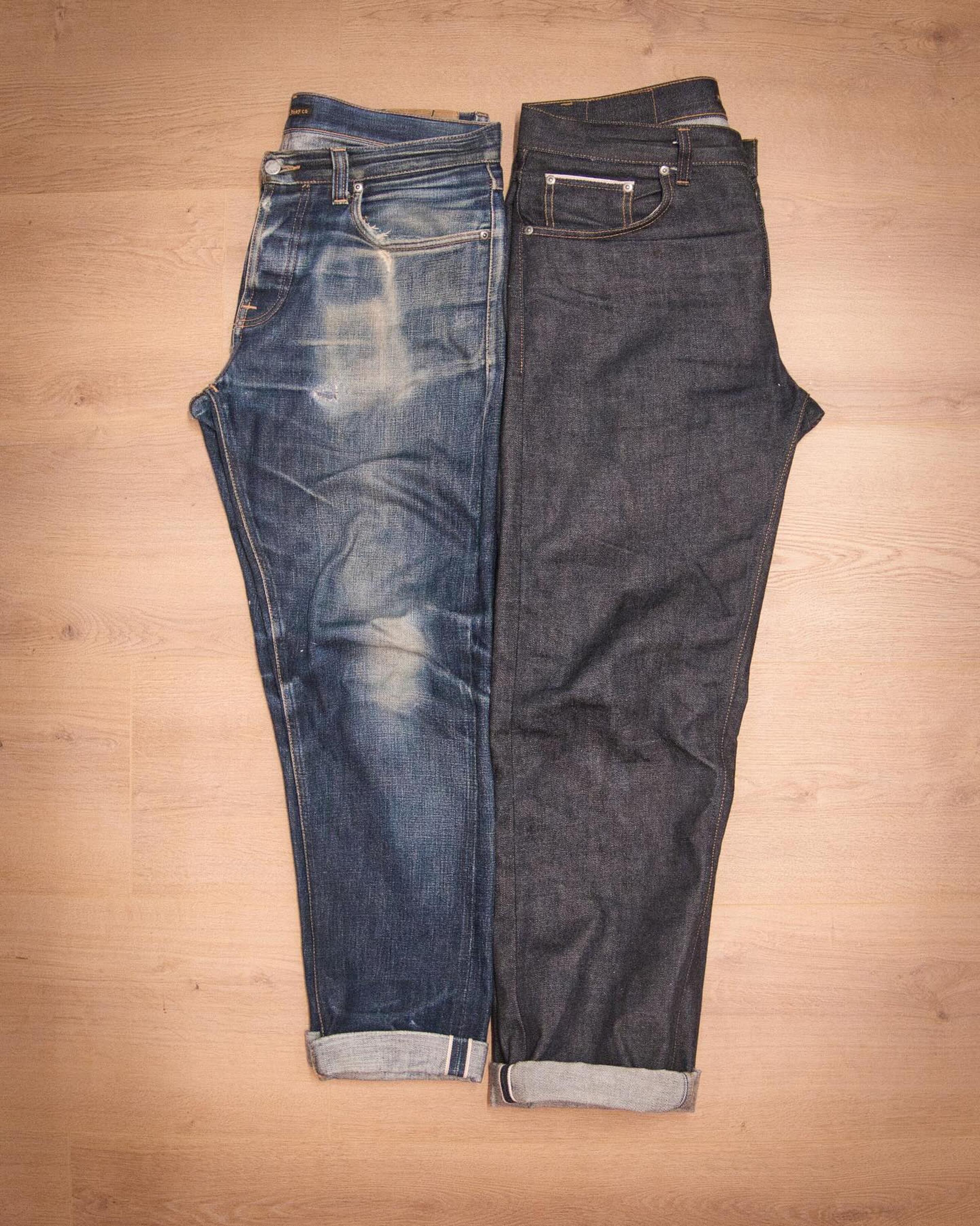 Two pairs of Nudie jeans lie side by side on a wooden floor, one faded and distressed, the other dark and new, both with rolled cuffs.