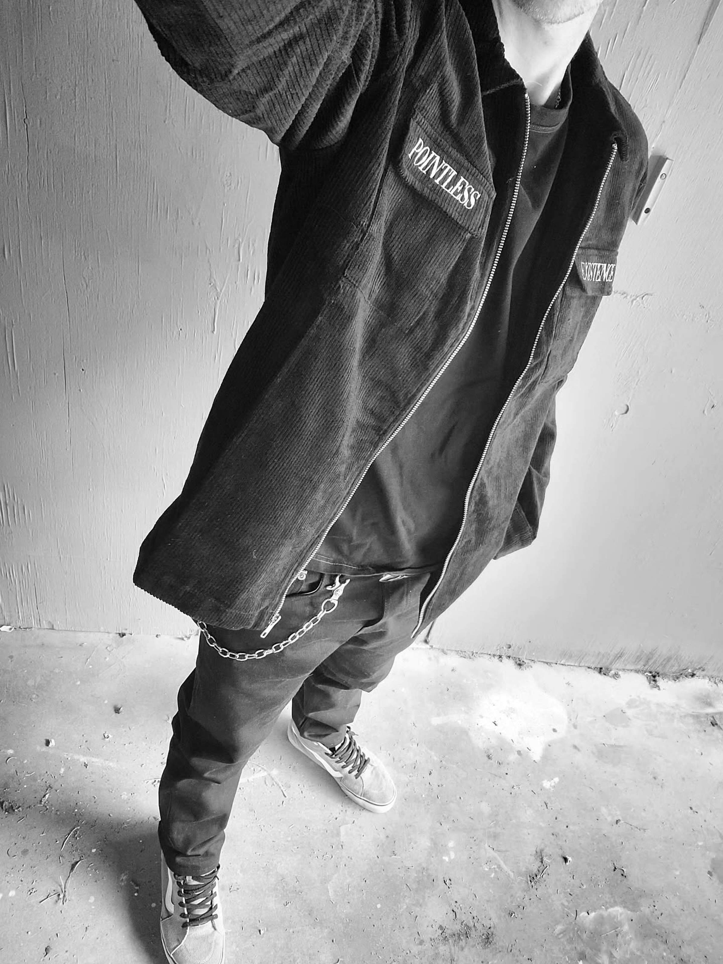 Person in black corduroy jacket and pants with chain accessory, standing on a concrete floor. Image is in black and white.