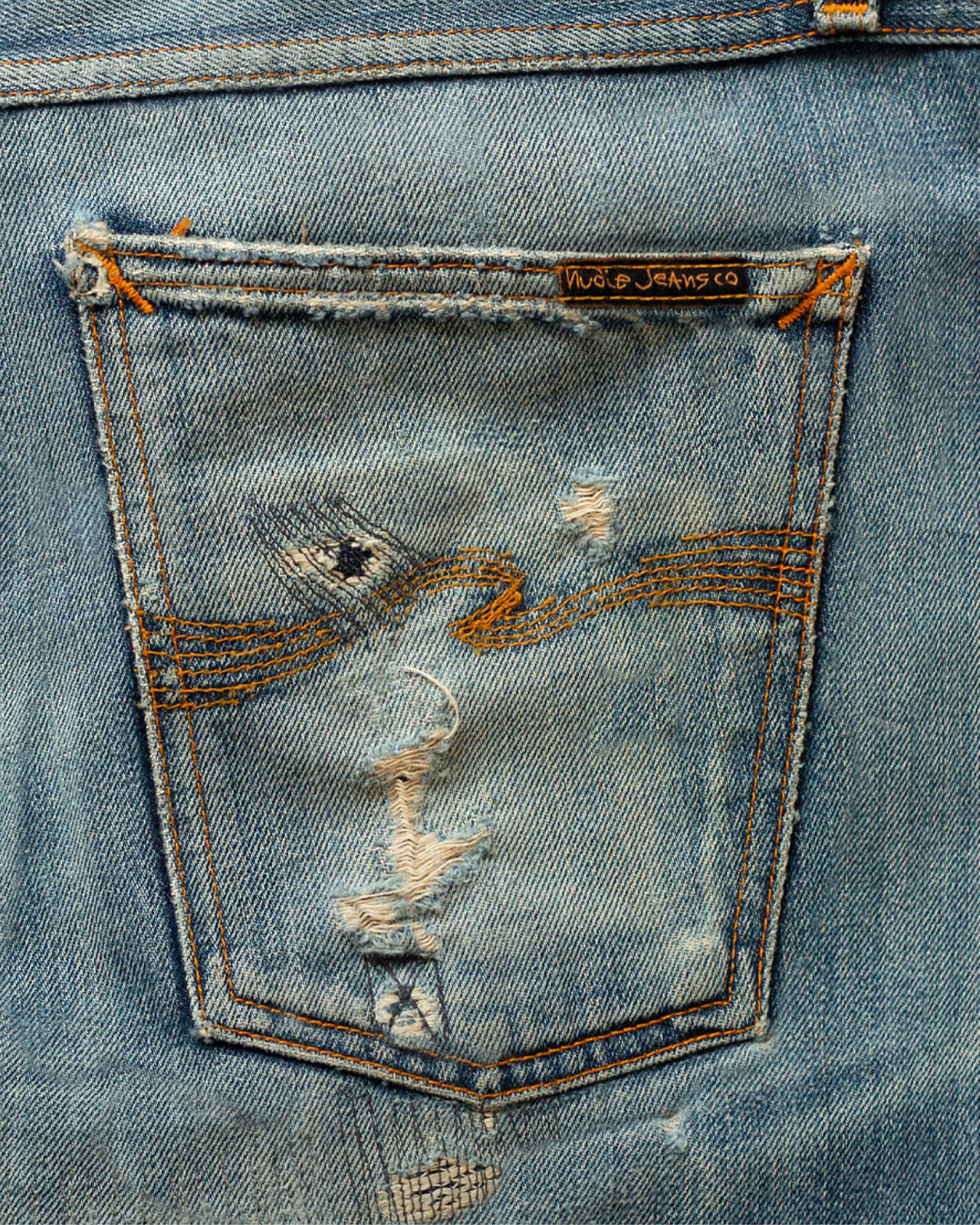 Close-up of a distressed Nudie Jeans pocket with visible rips and frayed edges, featuring orange stitching