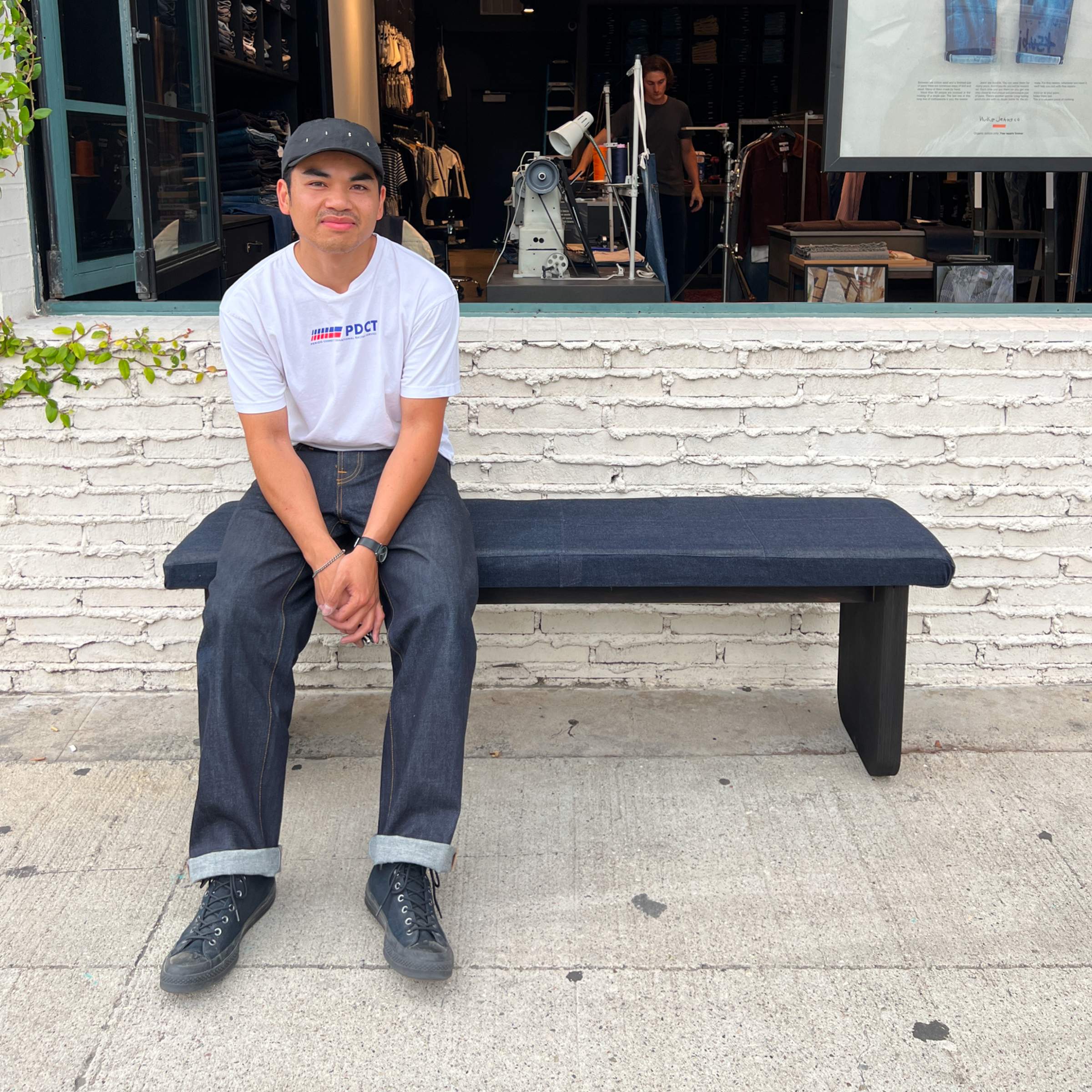 Paolo Manlucu in a white T-shirt and dark jeans sits on a bench outside a store with a brick wall background.