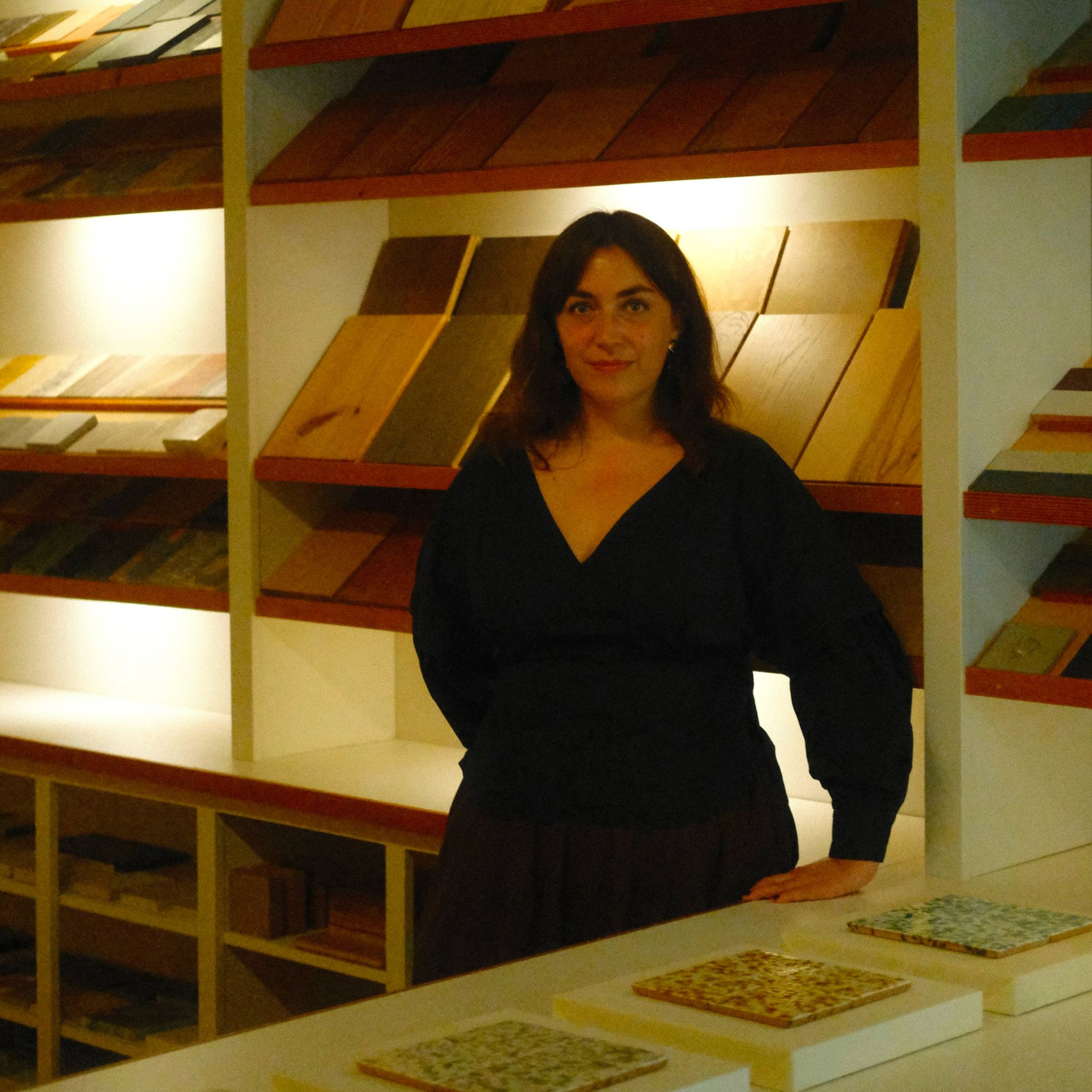 Emma Harling stands in a dimly lit showroom, surrounded by shelves displaying various tile samples.