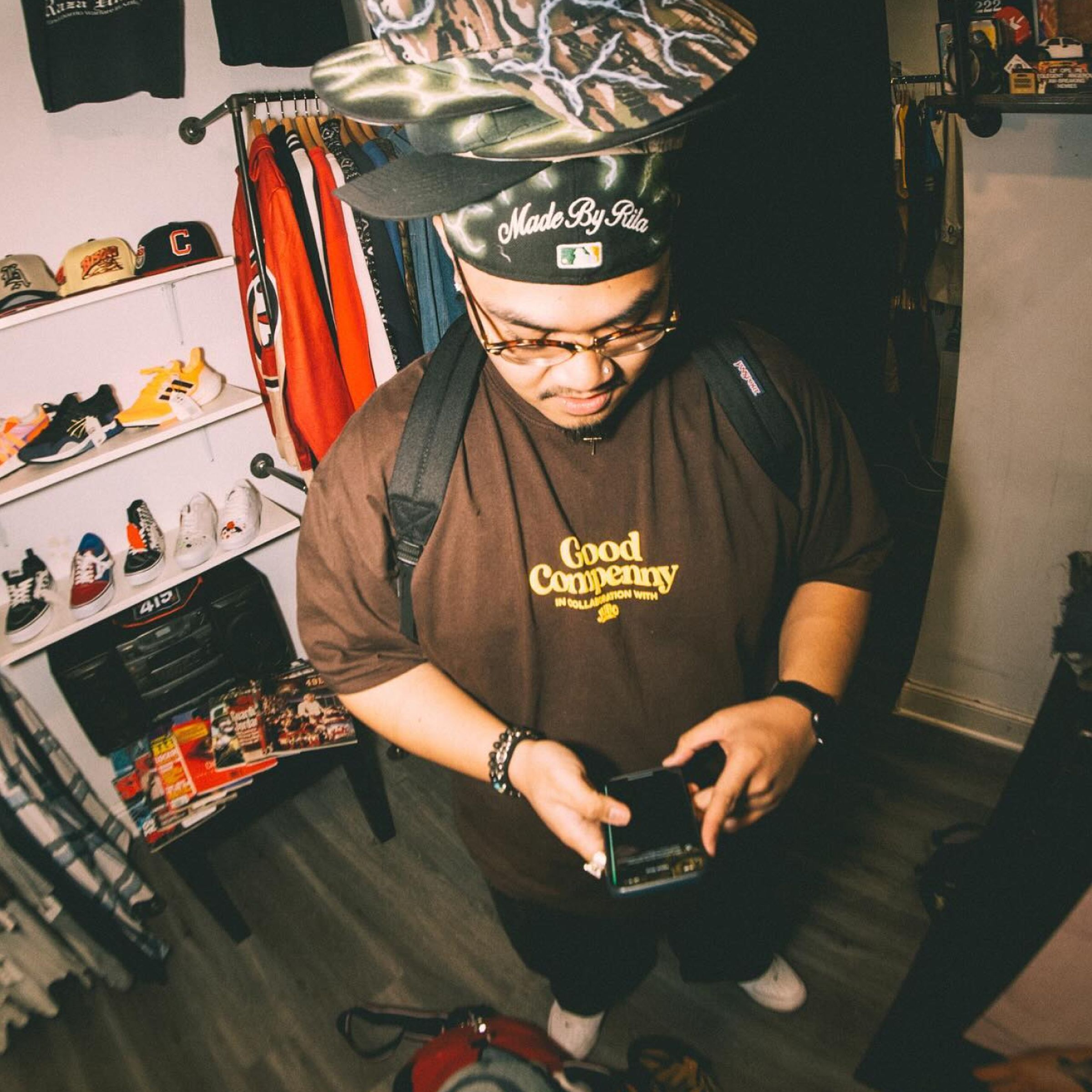Brian Nemendez in a brown "Good Company" t-shirt looking at a phone, surrounded by clothing and hats in a store setting.