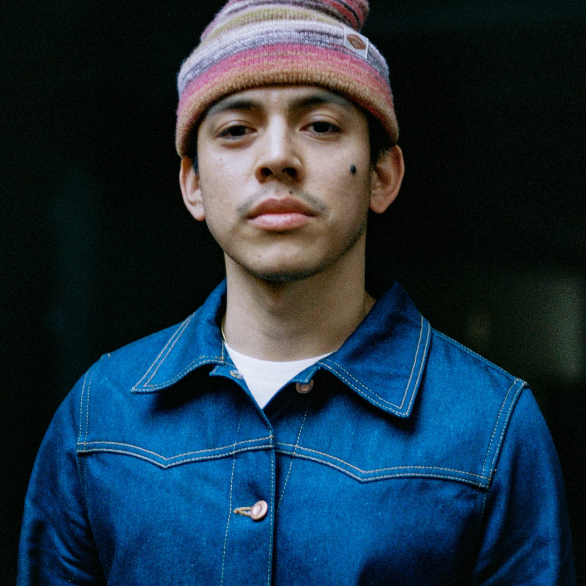 Josh from Nudie Jeans LA wearing a denim jacket and a multicolored beanie, standing against a dark background, looking at the camera.