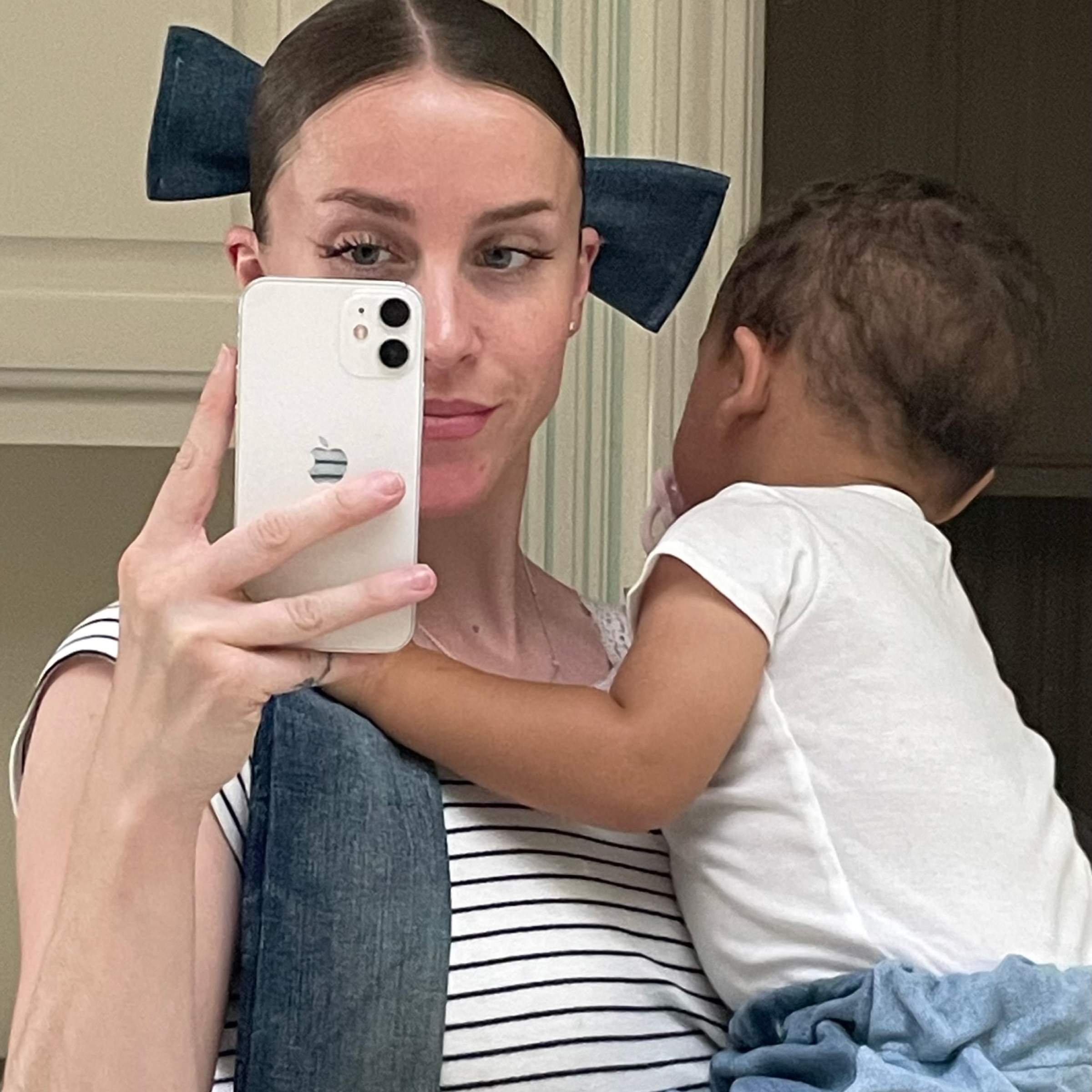 Alexandra Jenkins taking a mirror selfie holding a baby. She wears a large denim bow in her hair and a striped shirt. The baby wears a white shirt.