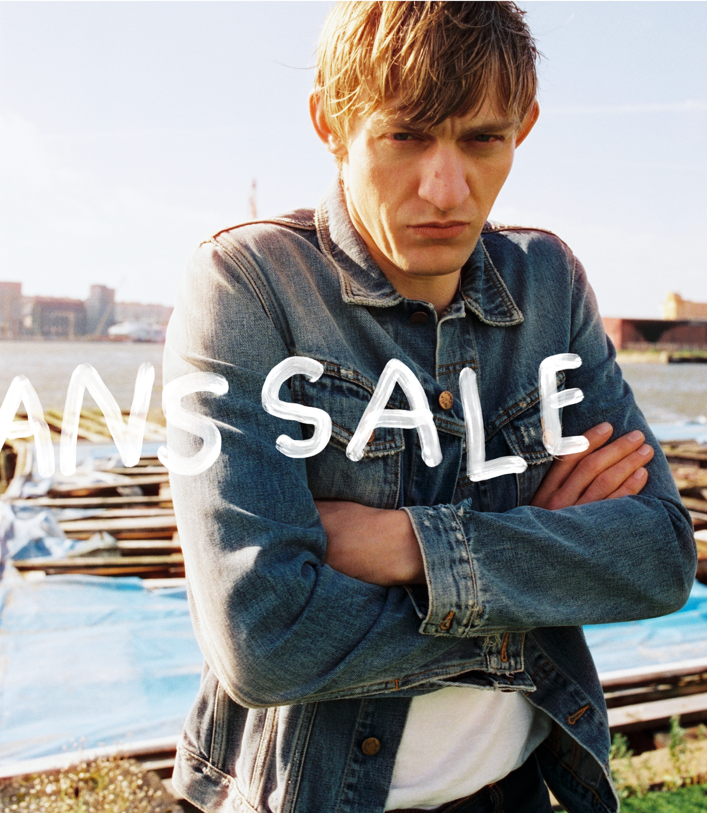 Man in denim jacket standing outdoors, arms crossed, with "JEANS SALE" text overlay. Urban background with bright sky.