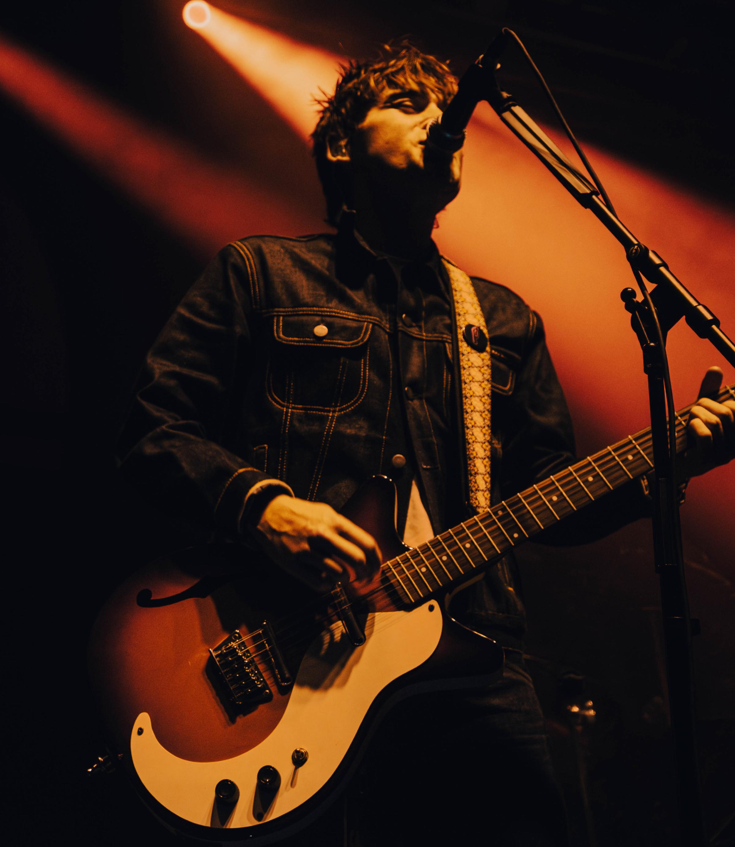 Guitarist in denim jacket singing into a microphone, playing electric guitar under warm orange stage lights.