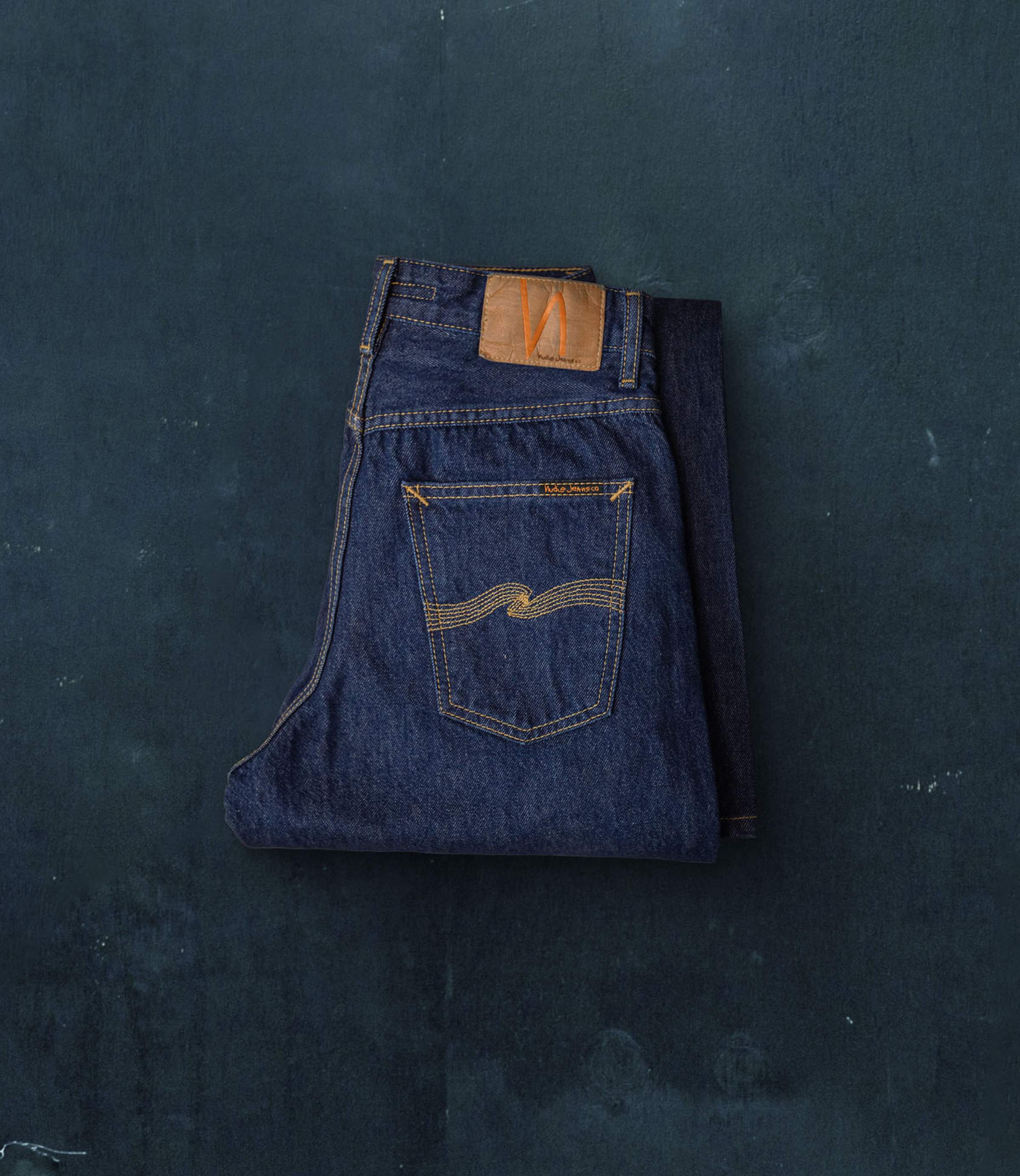 Folded pair of dark blue Nudie Jeans with a brown leather patch and yellow stitching on the back pocket, placed on a textured dark surface.