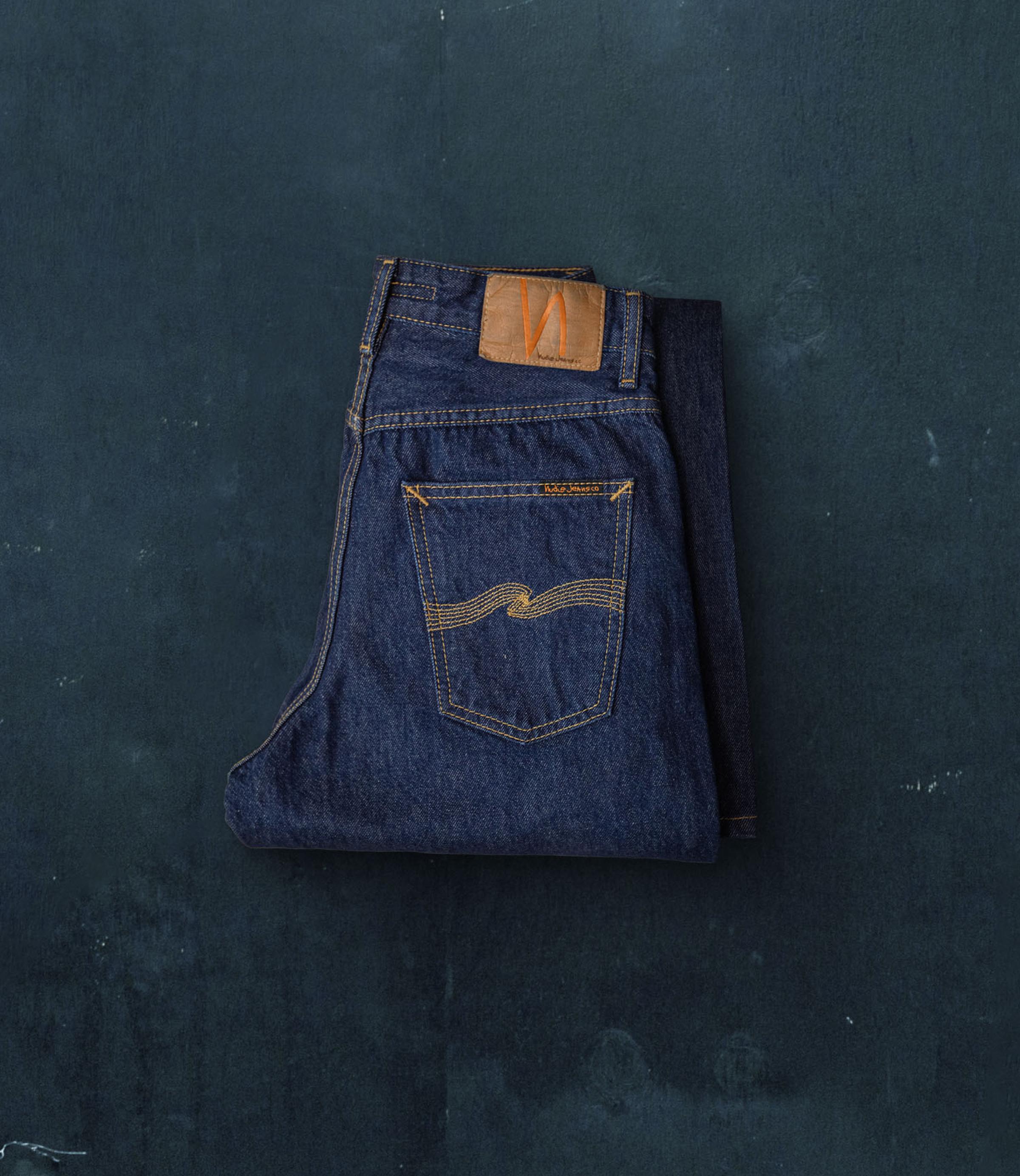 Folded pair of dark blue Nudie Jeans with a brown leather patch and yellow stitching on the back pocket, placed on a textured dark surface.