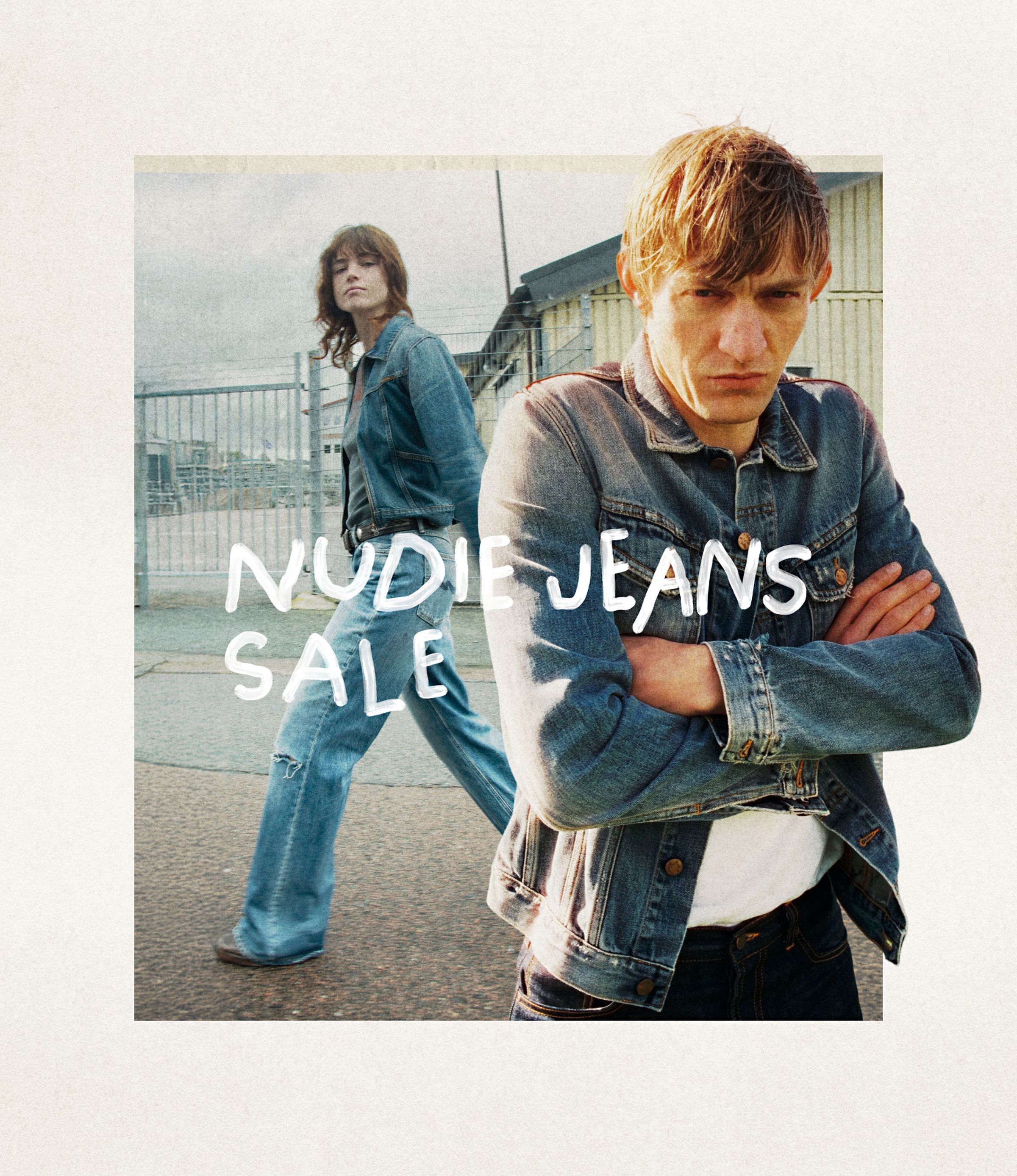 Two people in denim outfits; one stands with arms crossed, the other walks by. Text overlay reads "Nudie Jeans Sale." Industrial background.