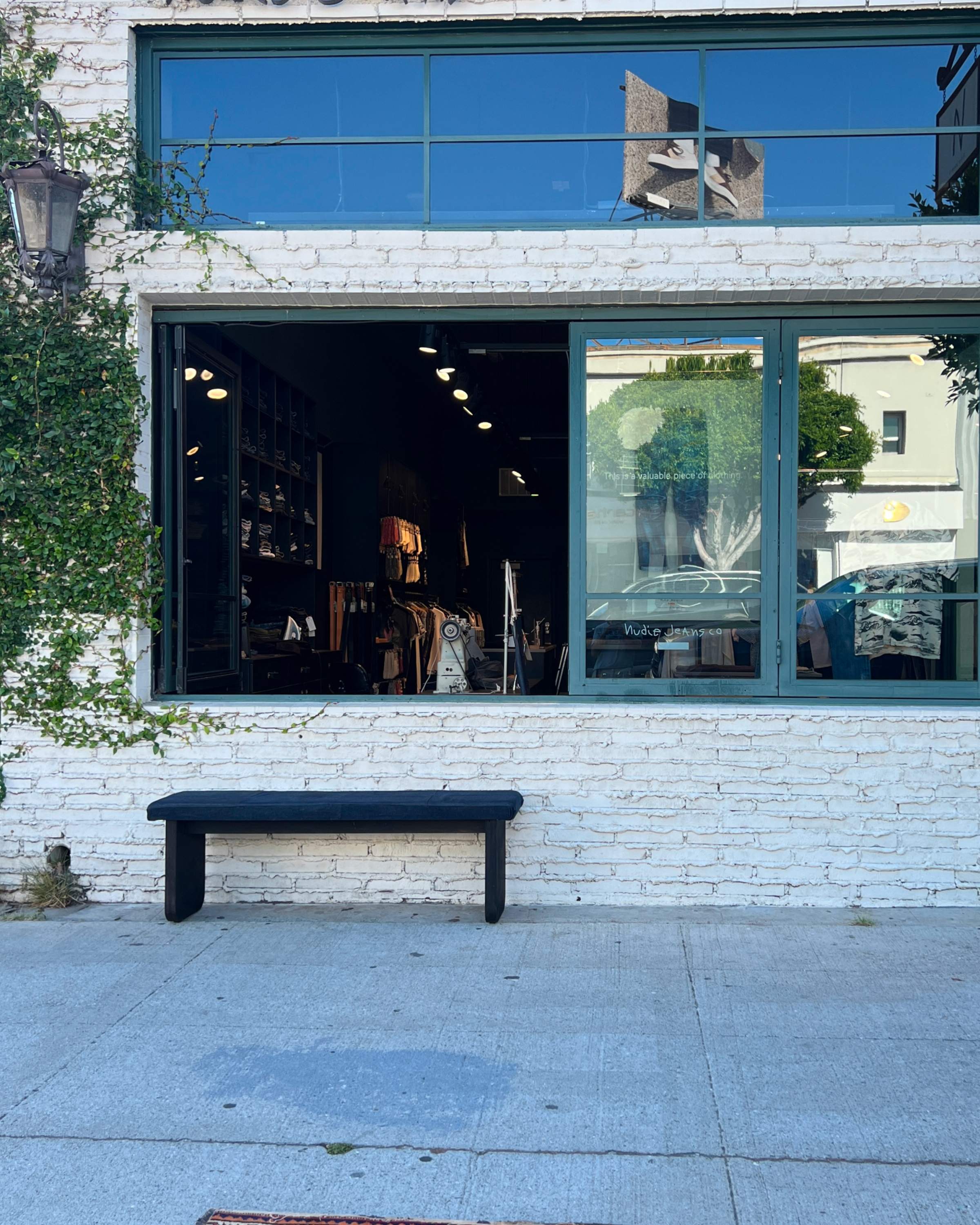 A brick storefront with a large window displaying interior items. A black bench is placed on the sidewalk outside. Green ivy climbs the wall on the left.
