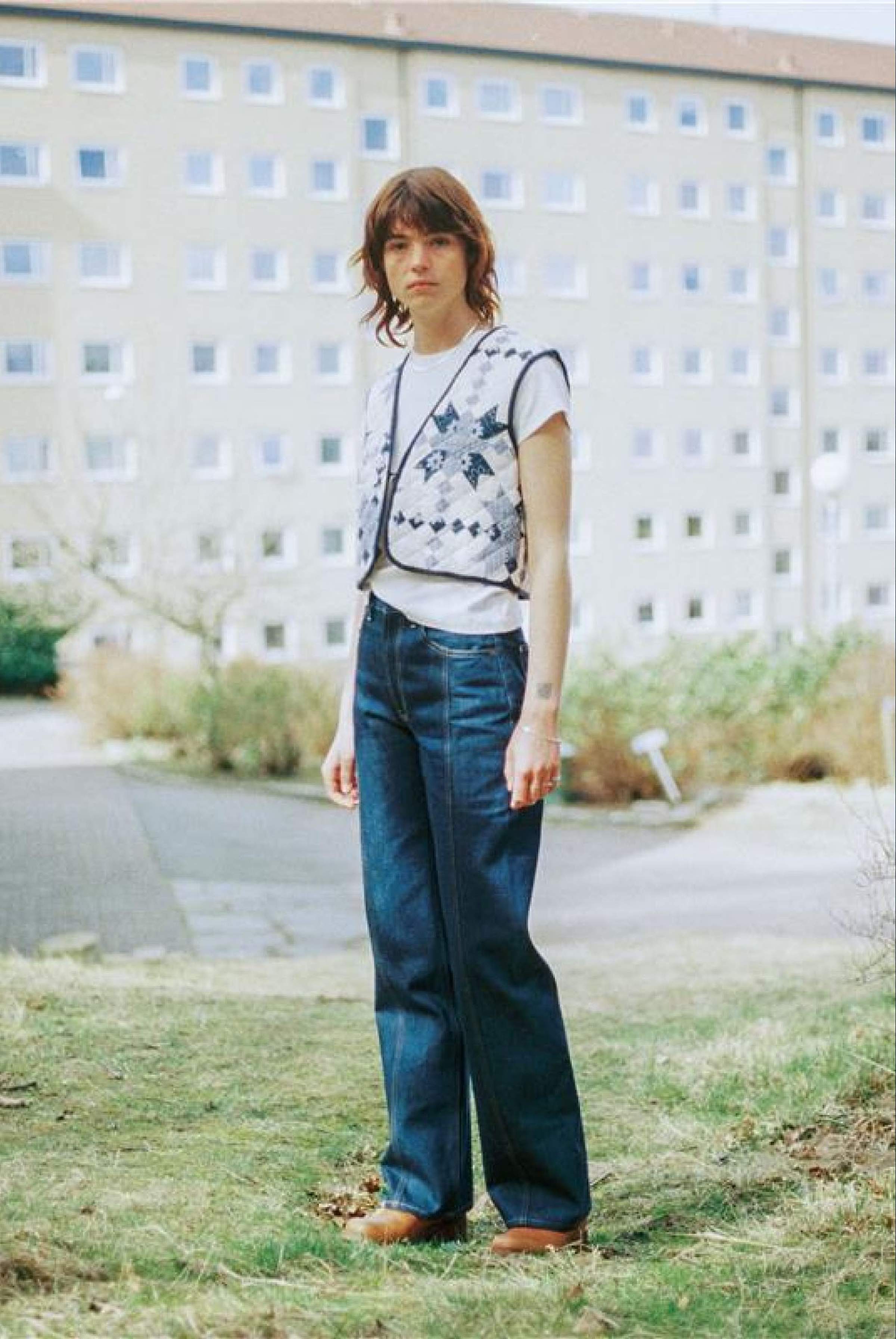 Person standing on grass wearing a white shirt, patterned vest, and dry wide-leg jeans, with a building in the background.