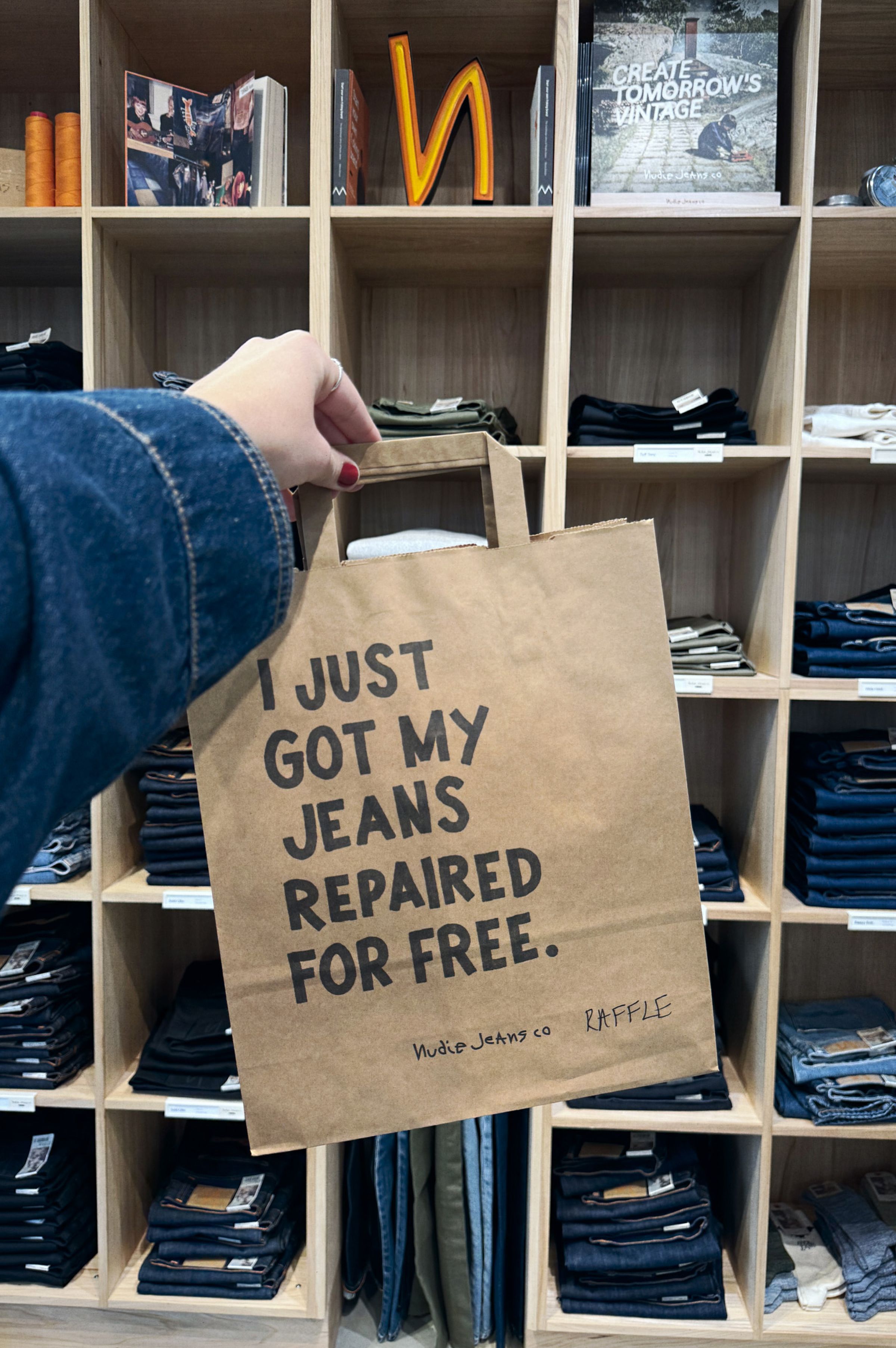 A hand holds a paper bag labeled "I just got my jeans repaired for free" in front of shelves filled with folded denim jeans.