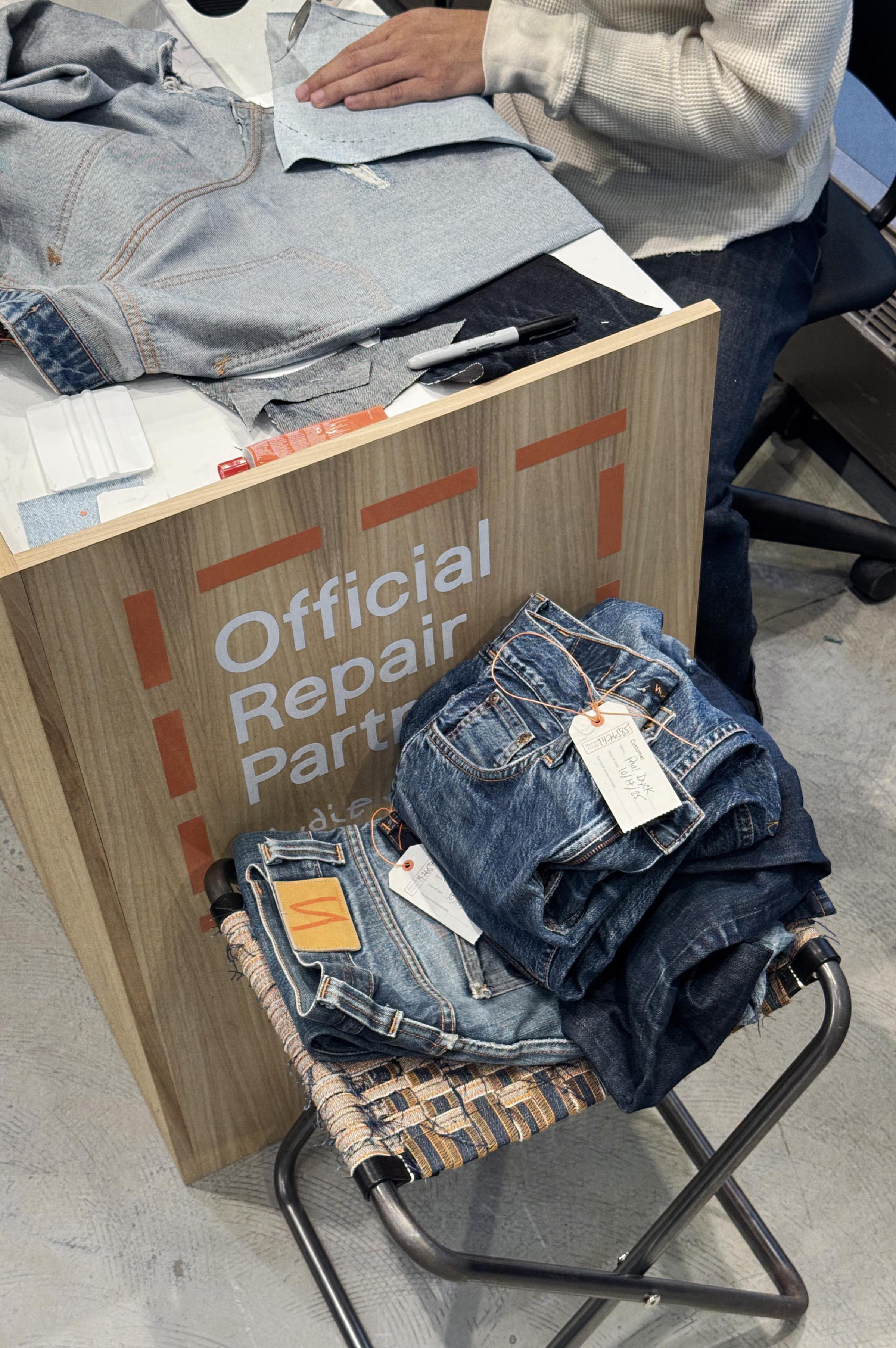 Person repairing denim at a wooden desk labeled "Official Repair Partner," with a stack of jeans on a nearby woven stool.