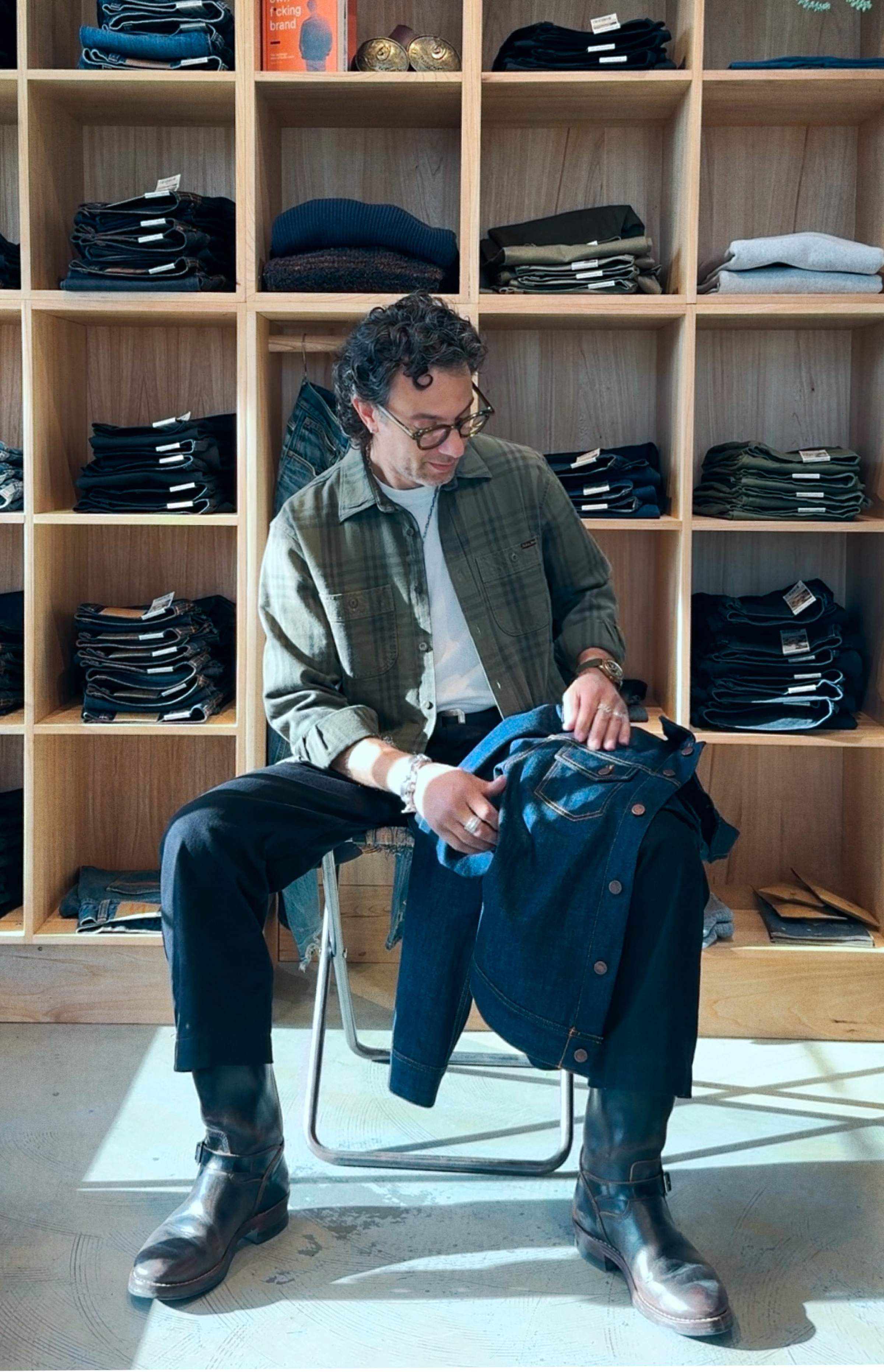 Person sitting in a clothing store, holding denim, surrounded by wooden shelves filled with folded clothes.