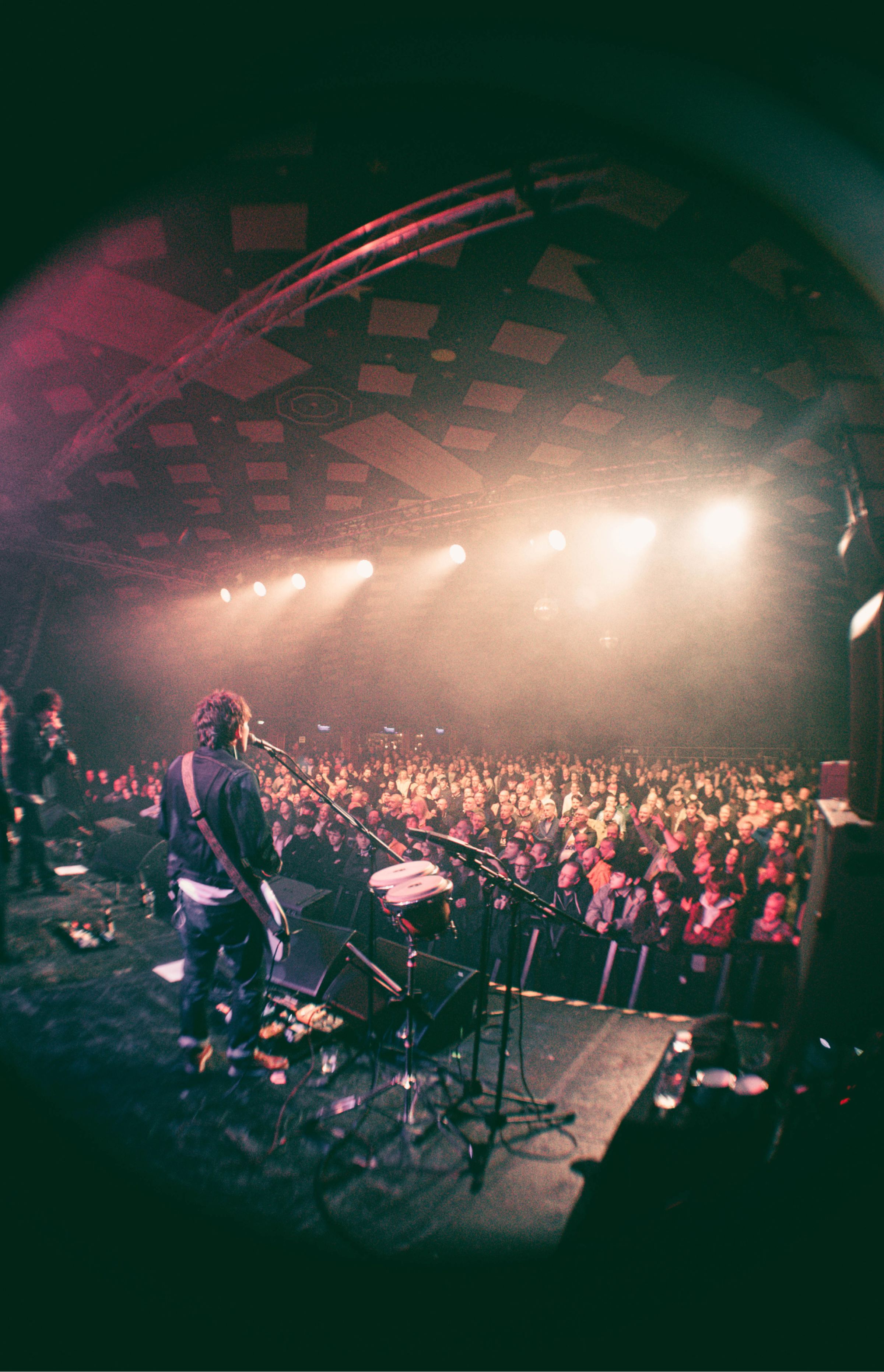 Pastel on stage seen from behind singing into a microphone as a packed, illuminated crowd watches under hazy stage lights.