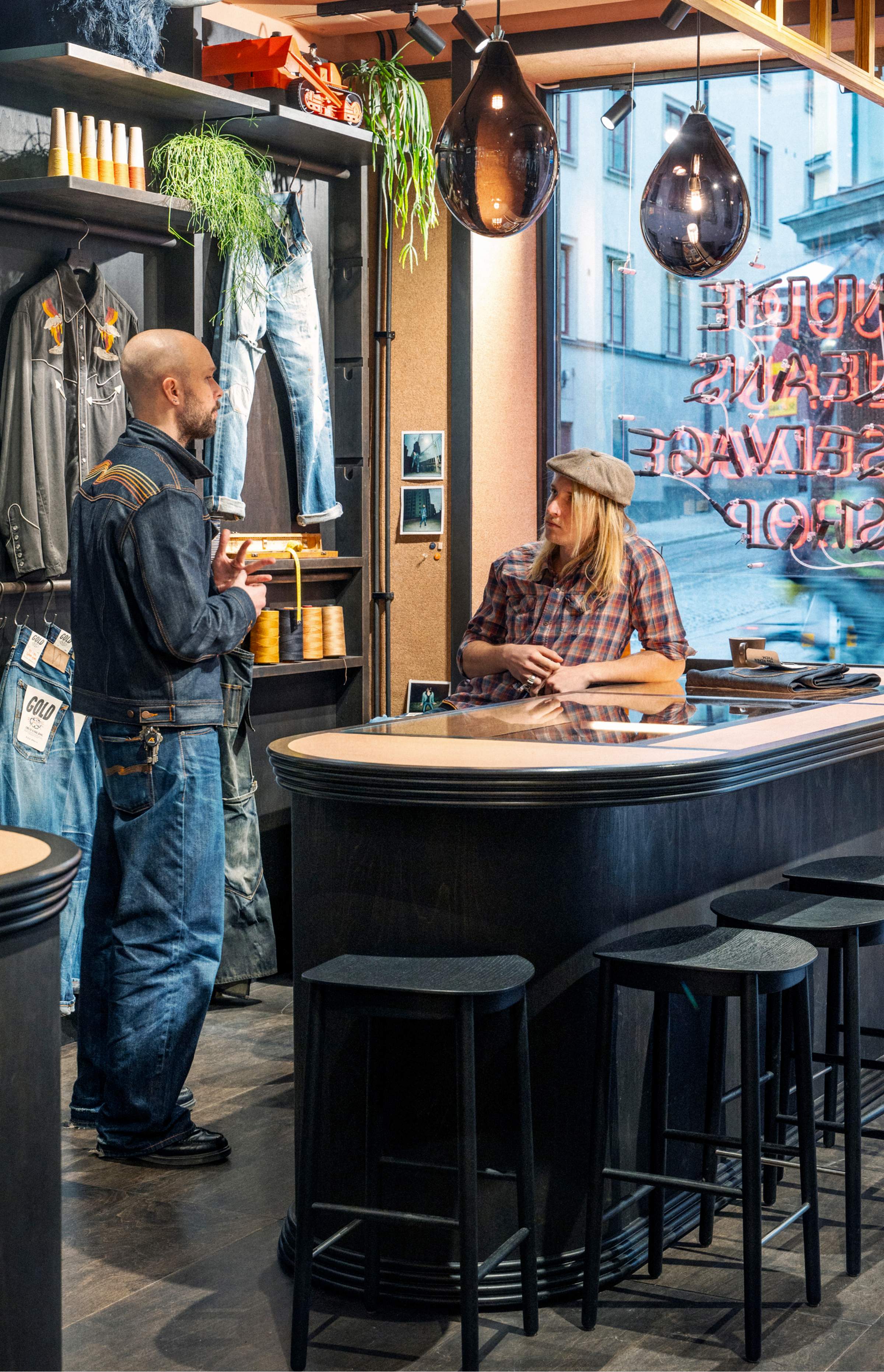 Two people in a denim-themed store, one in a leather jacket and jeans, the other in casual attire, engaging in conversation near a counter.