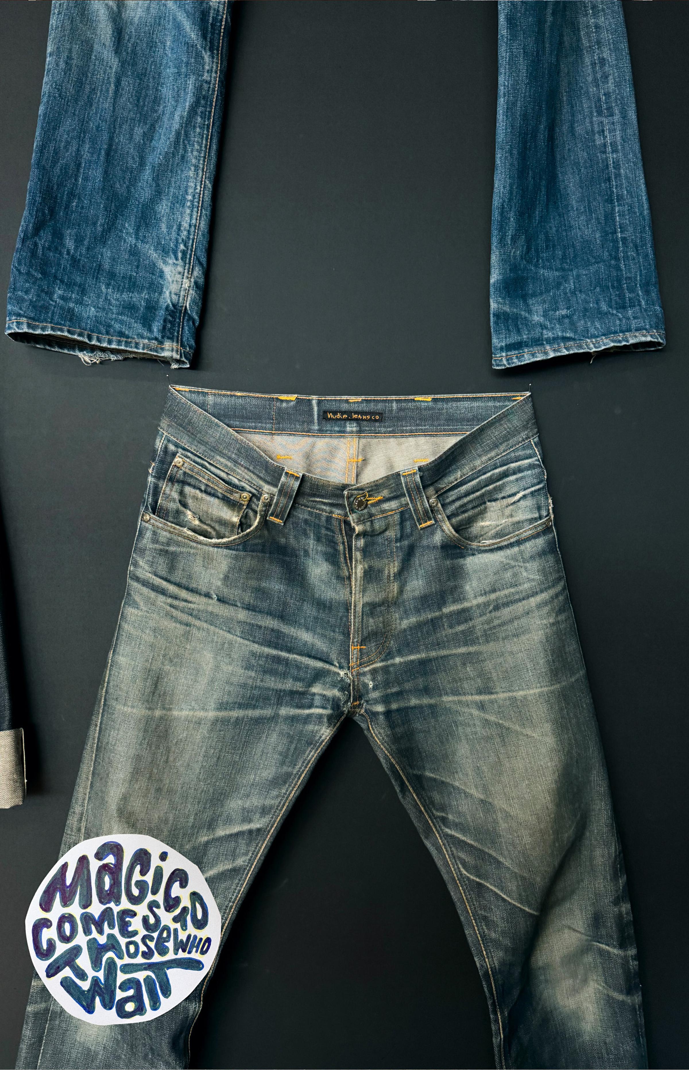 A pair of worn blue jeans arranged in an upside-down "T" shape on a black background with a text bubble saying, "Magic Comes To Those Who Wait."