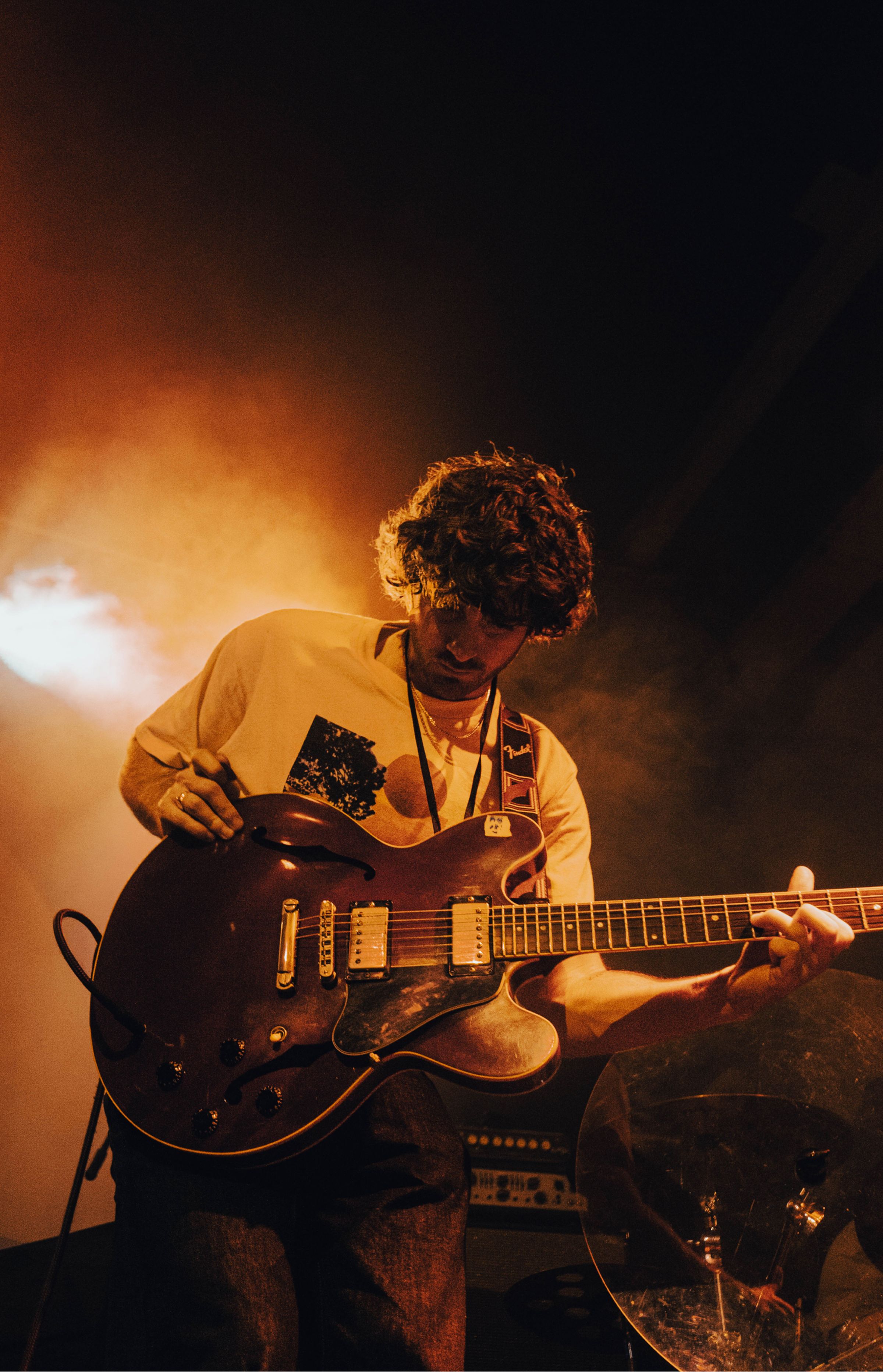 Musician with curly hair looking down, playing a hollow-body electric guitar on stage under warm orange lighting.