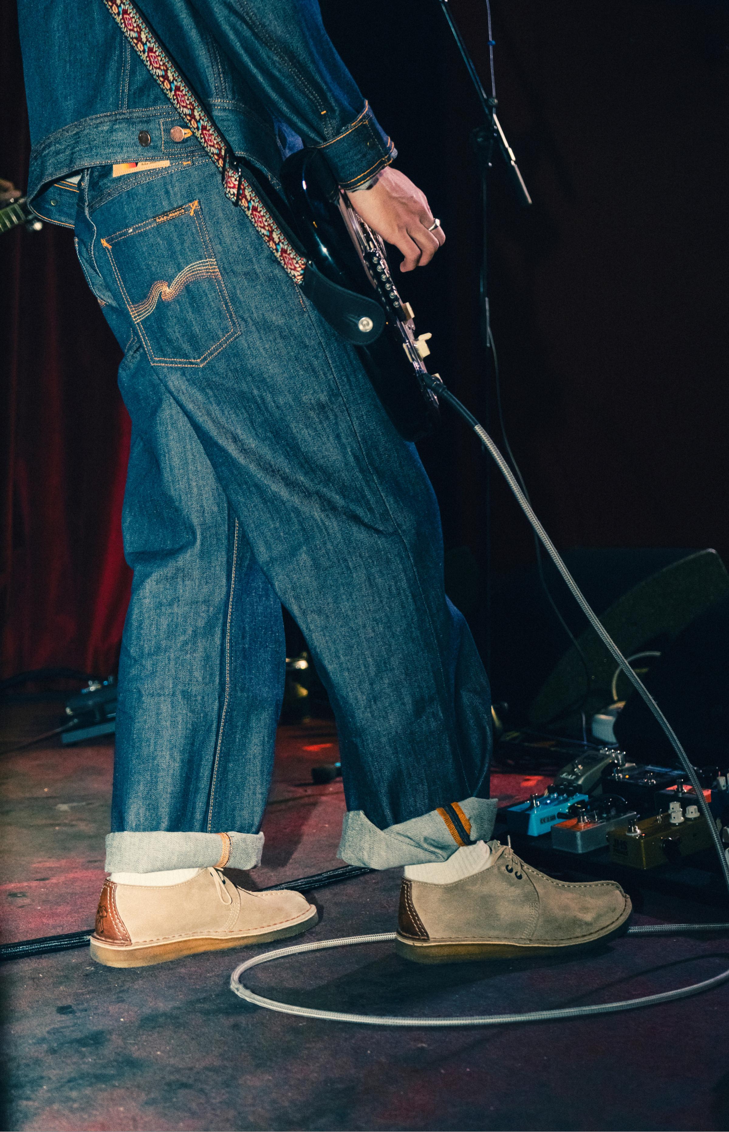 Lower body of a guitarist on stage: rolled upp Tuff Tony Sunburns Nudie Jeans, suede shoes, patterned strap, electric guitar, pedals and cables.