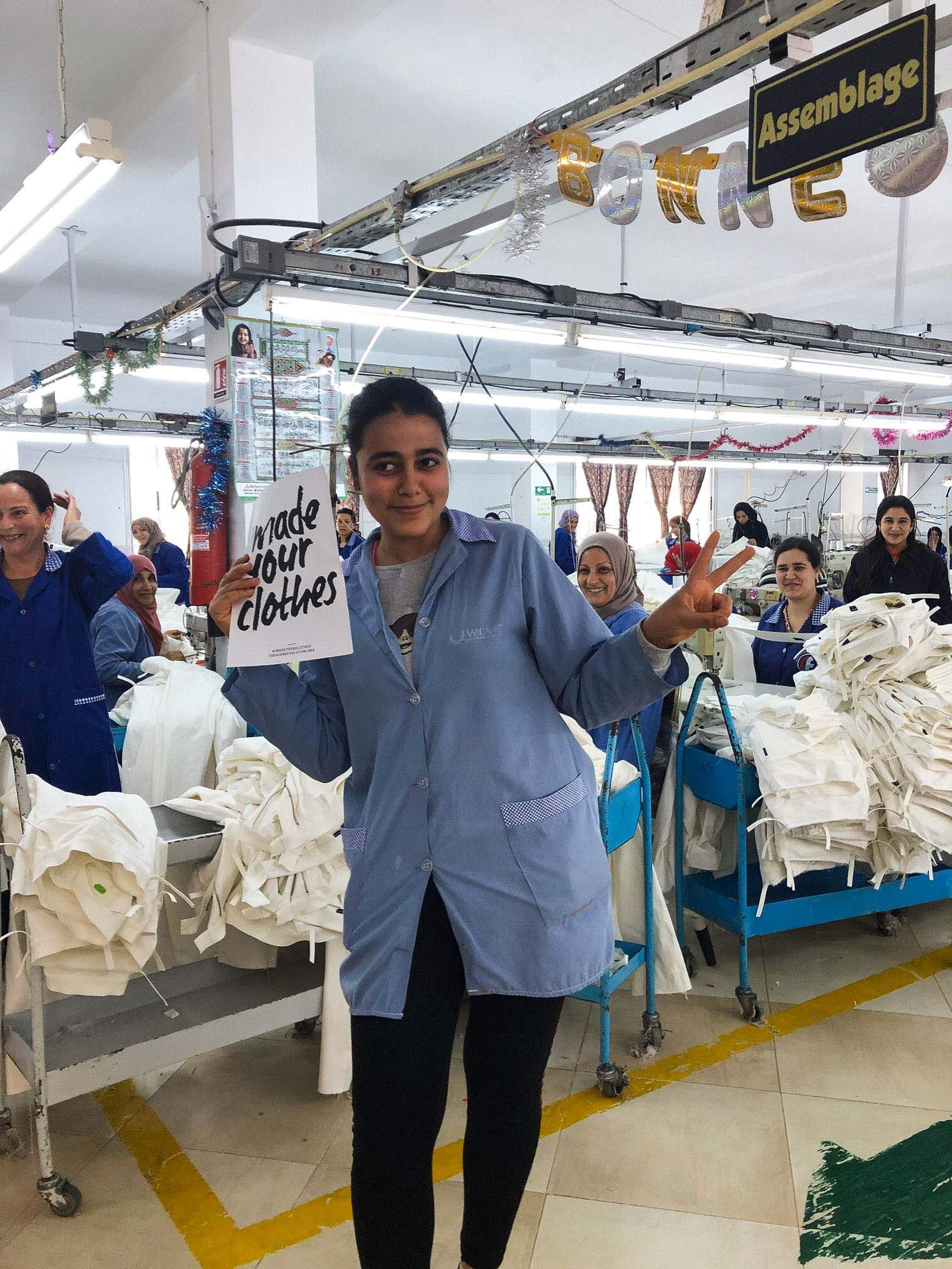 A woman in a factory setting holds a sign reading "I made your clothes," surrounded by coworkers and fabric, under a sign labeled "Assemblage."