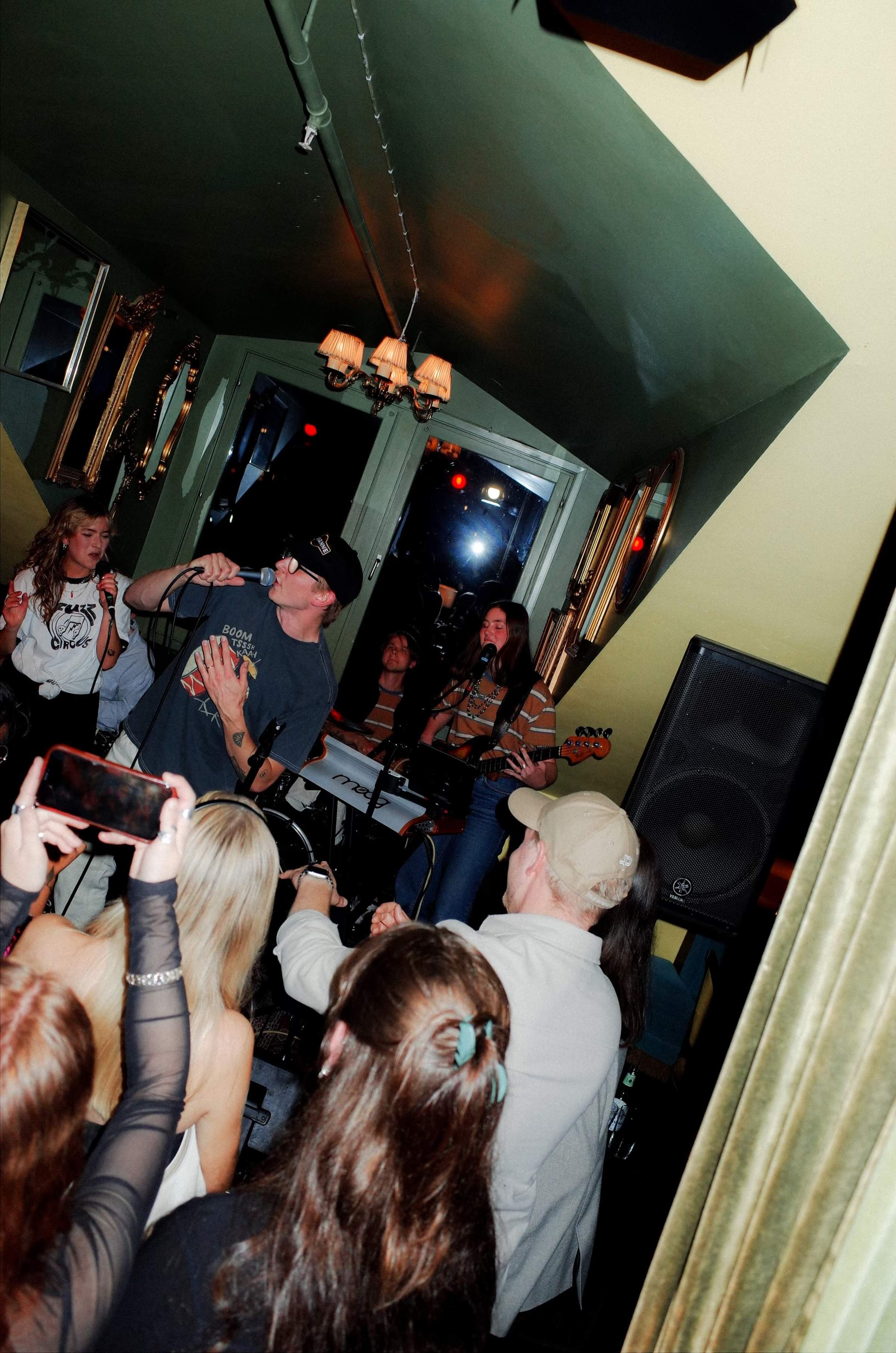 Mattis performs on a dimly lit stage with a musicians, and instruments. The audience's hands are visible in the foreground.