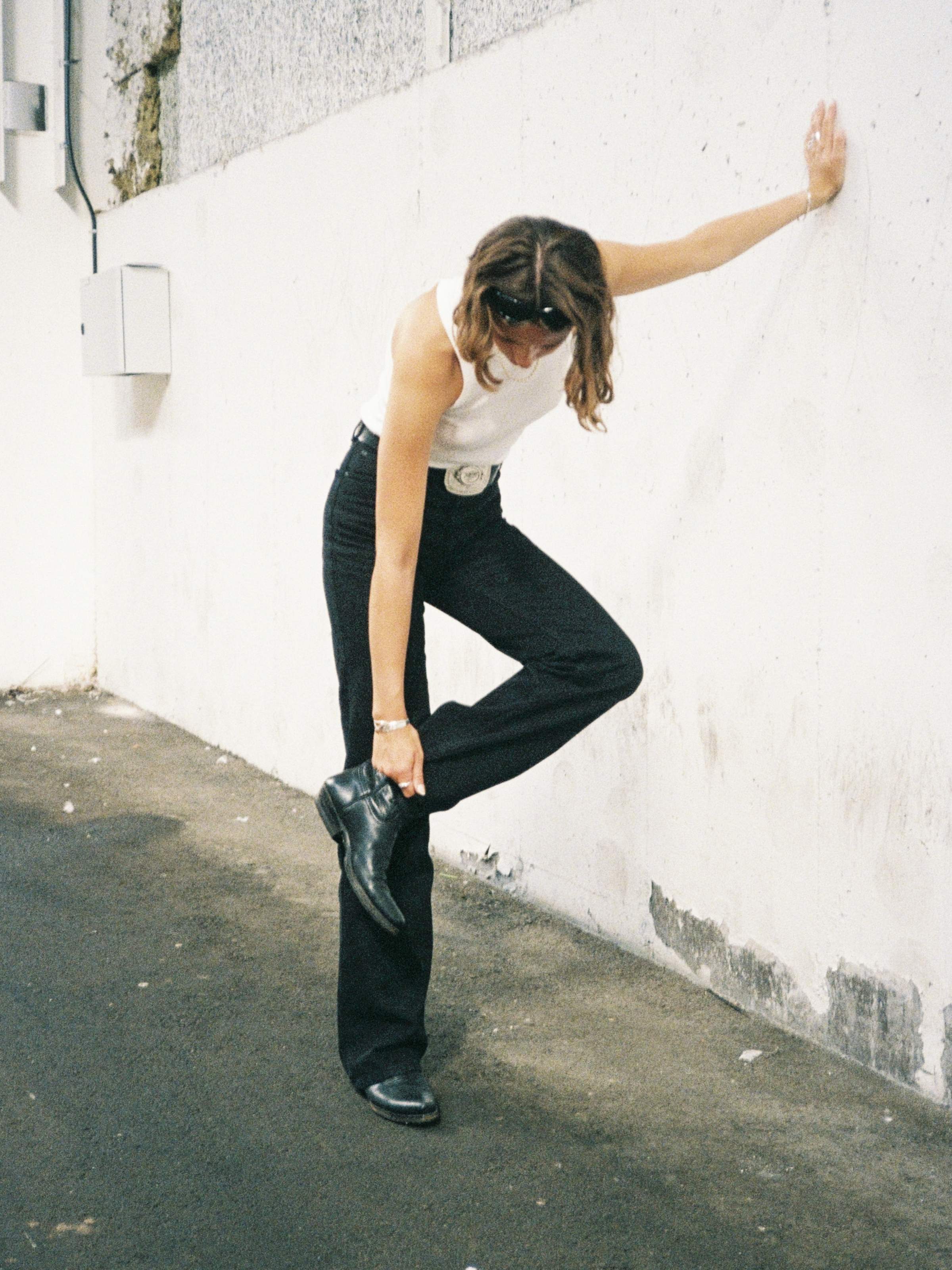 Women in a white tank top and black pants leans against a wall, adjusting their black boot on a textured urban street.