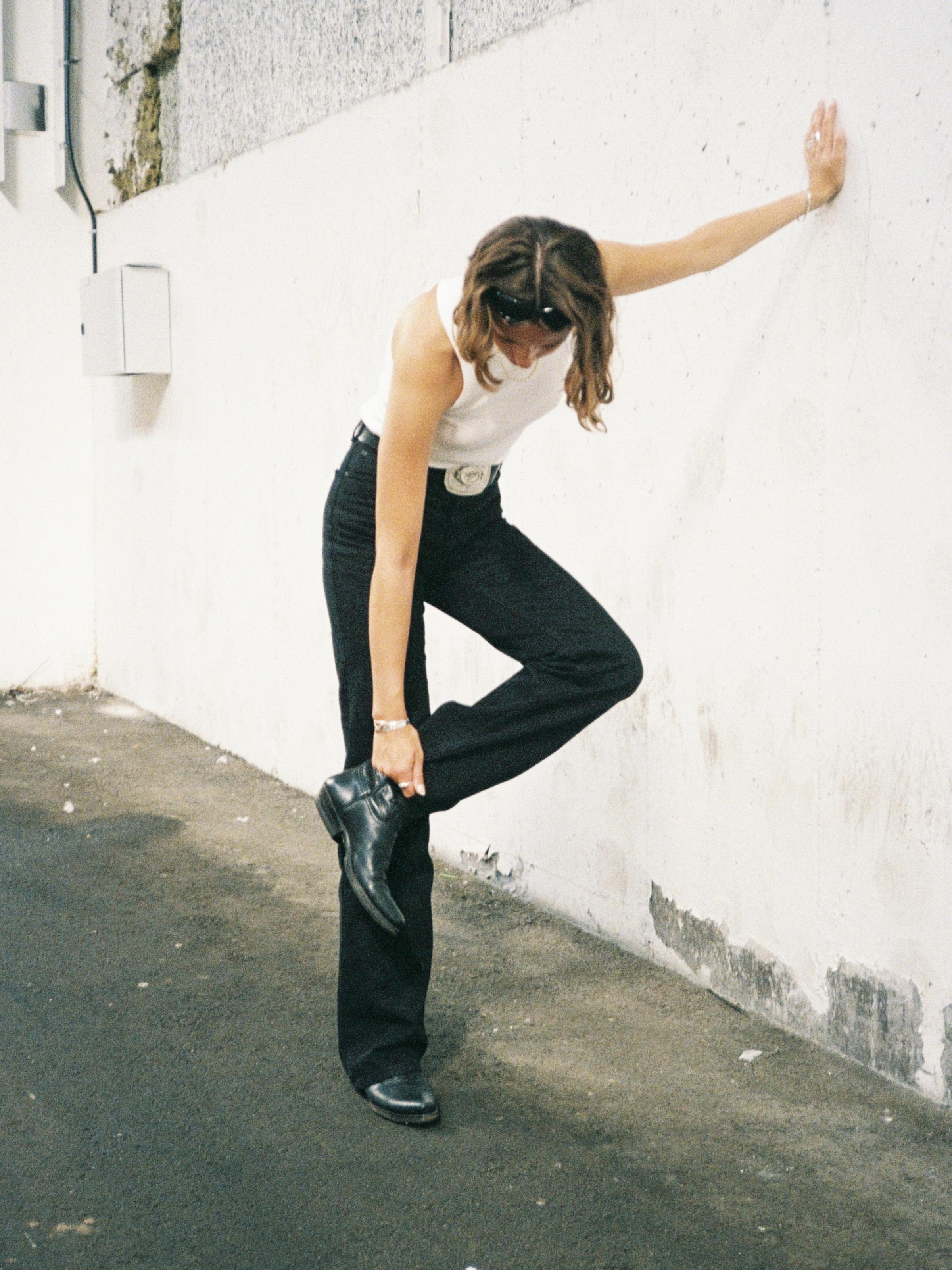 Women in a white tank top and black pants leans against a wall, adjusting their black boot on a textured urban street.