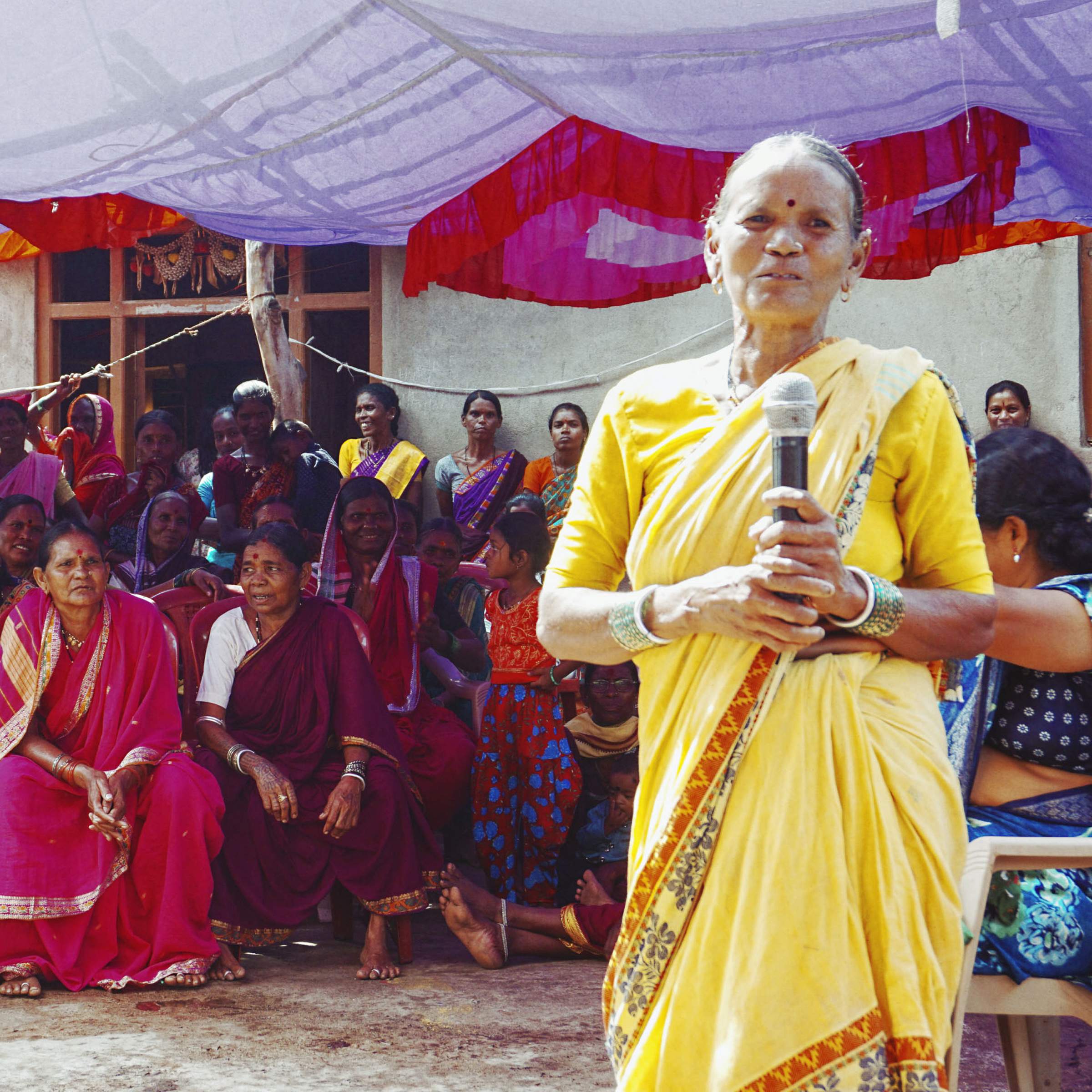 Elderly woman in a yellow sari speaks into a microphone in front of a seated group under a tent with colorful decorations.