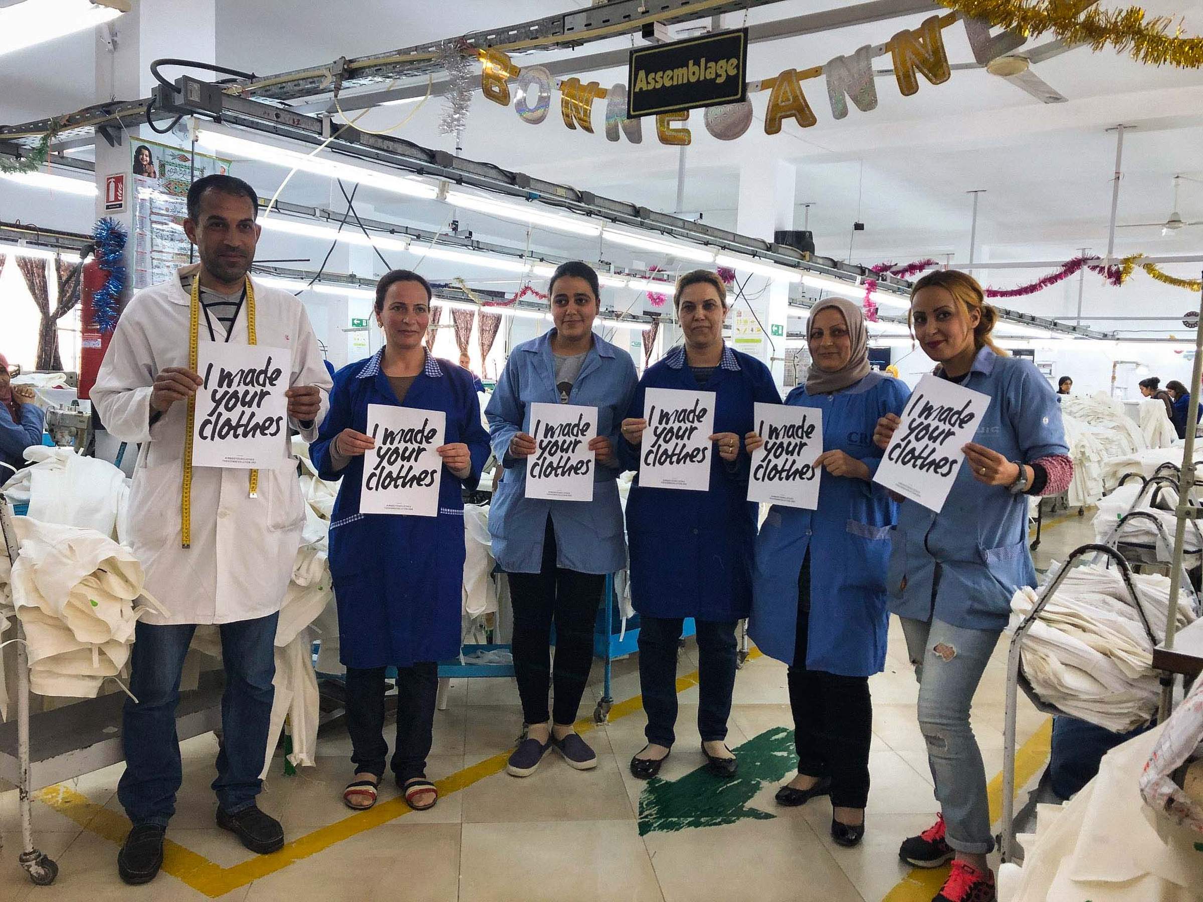 Factory workers in blue uniforms hold signs reading "I made your clothes," standing in a garment assembly area with festive decorations.