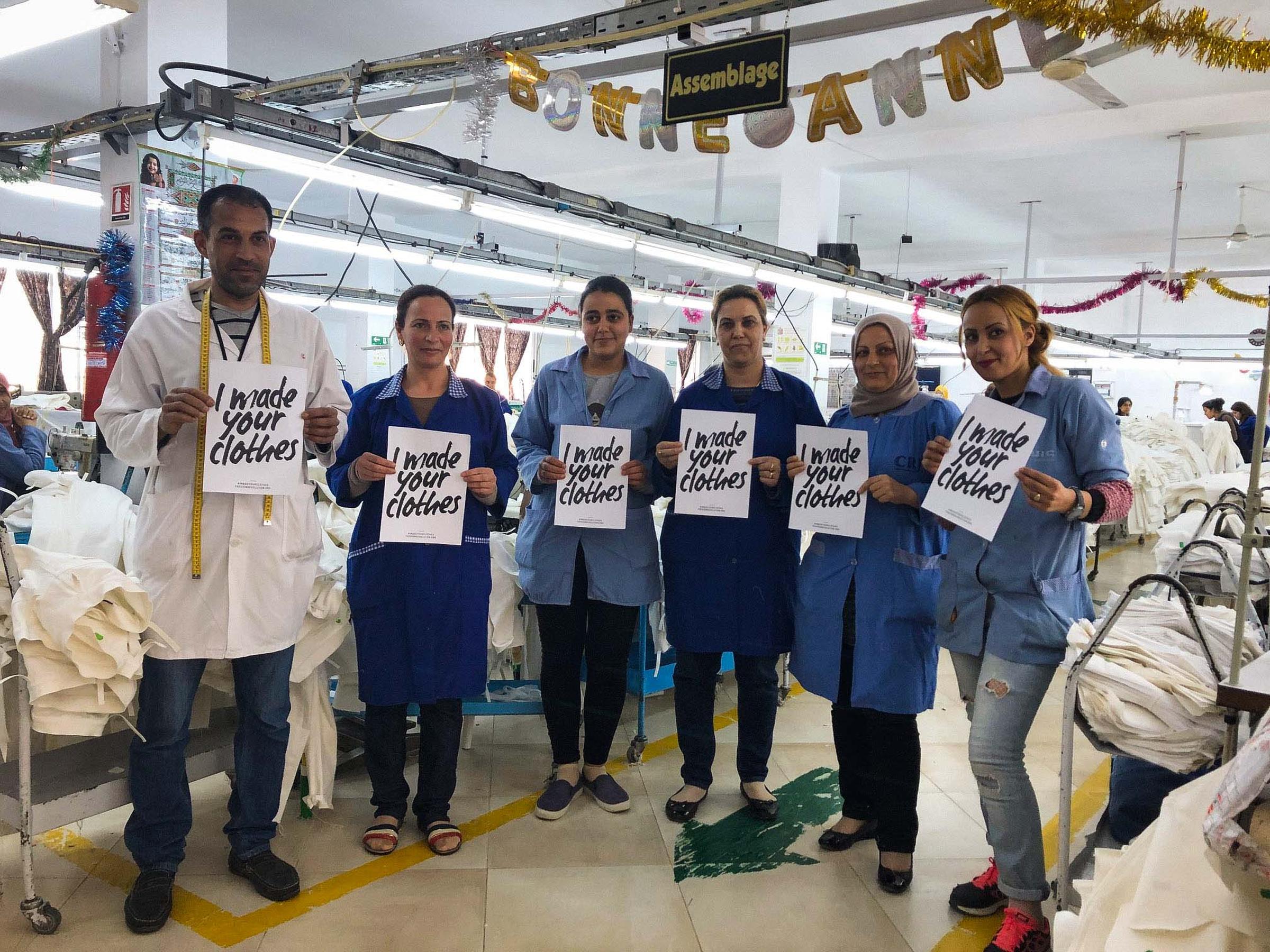 Factory workers in blue uniforms hold signs reading "I made your clothes," standing in a garment assembly area with festive decorations.