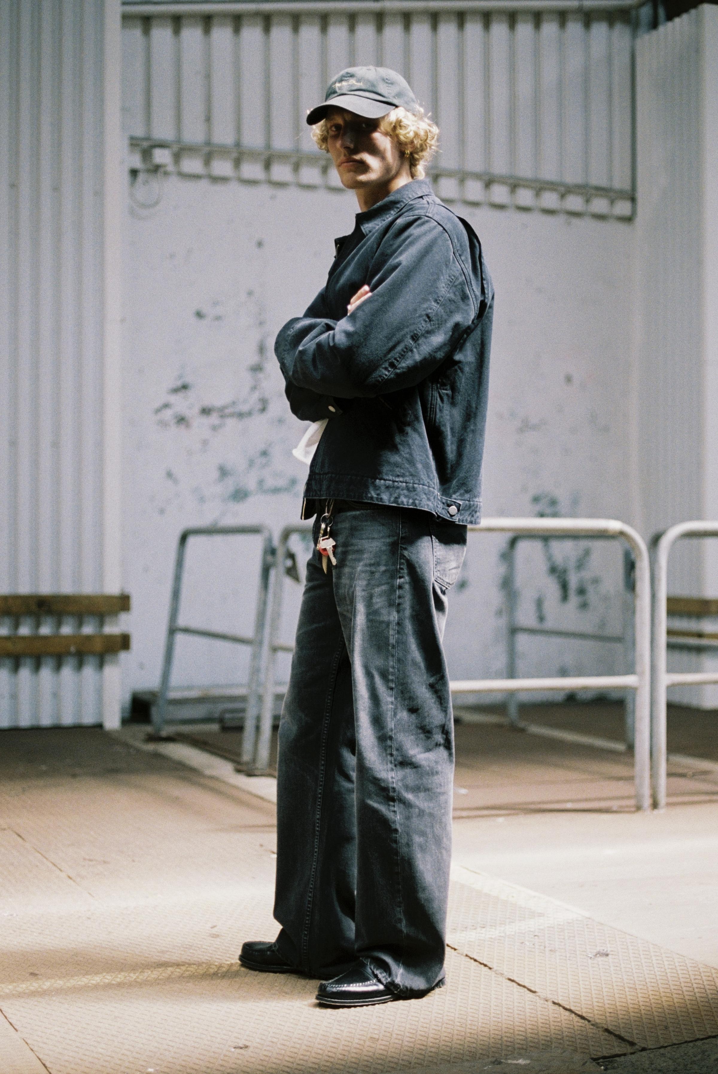 A man in black Loud Larry Wide Jeans and cap stands with arms crossed in an industrial setting, featuring metal railings and a textured wall.