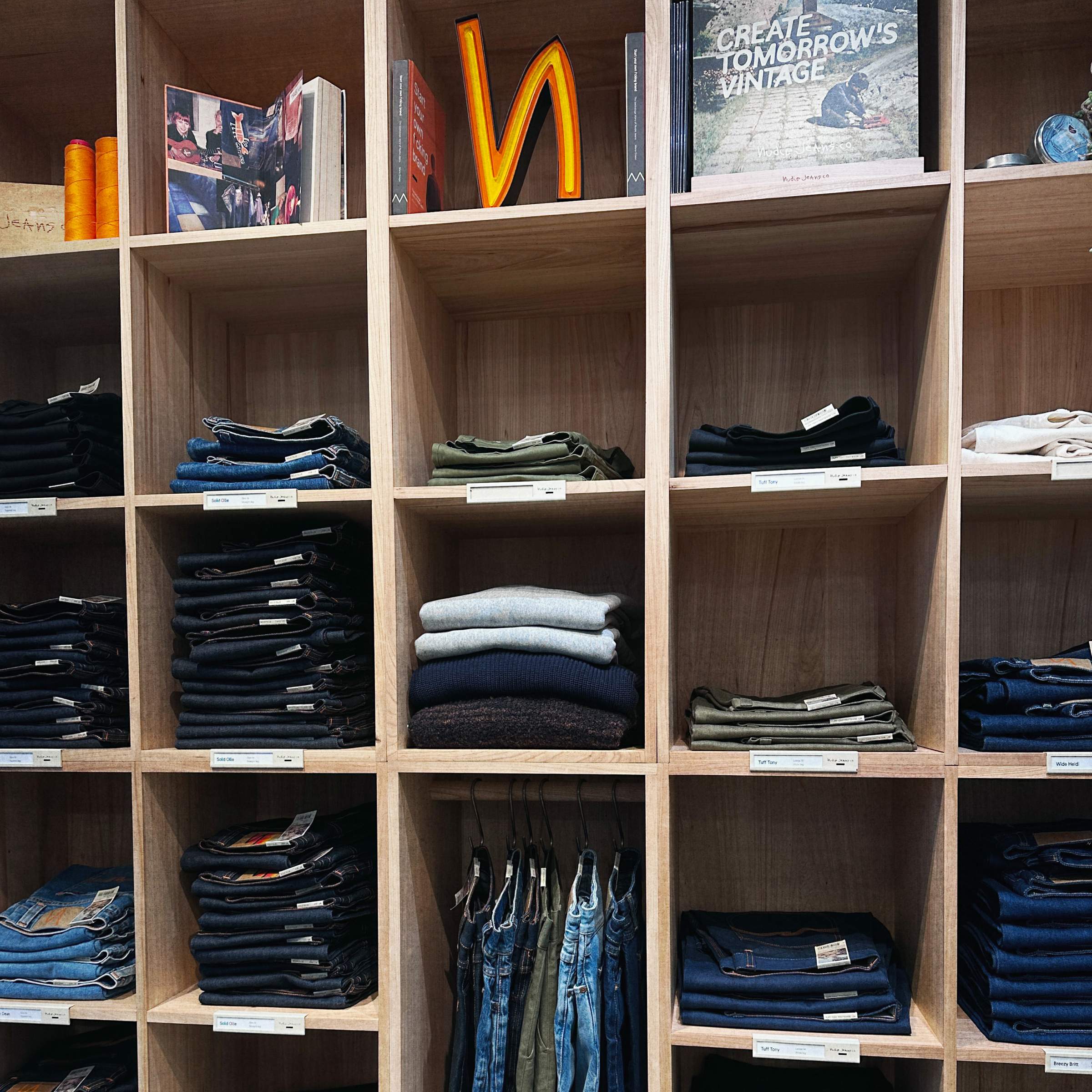 Neatly folded jeans and sweaters organized in wooden shelves with books and signs on top. Tags label each section.