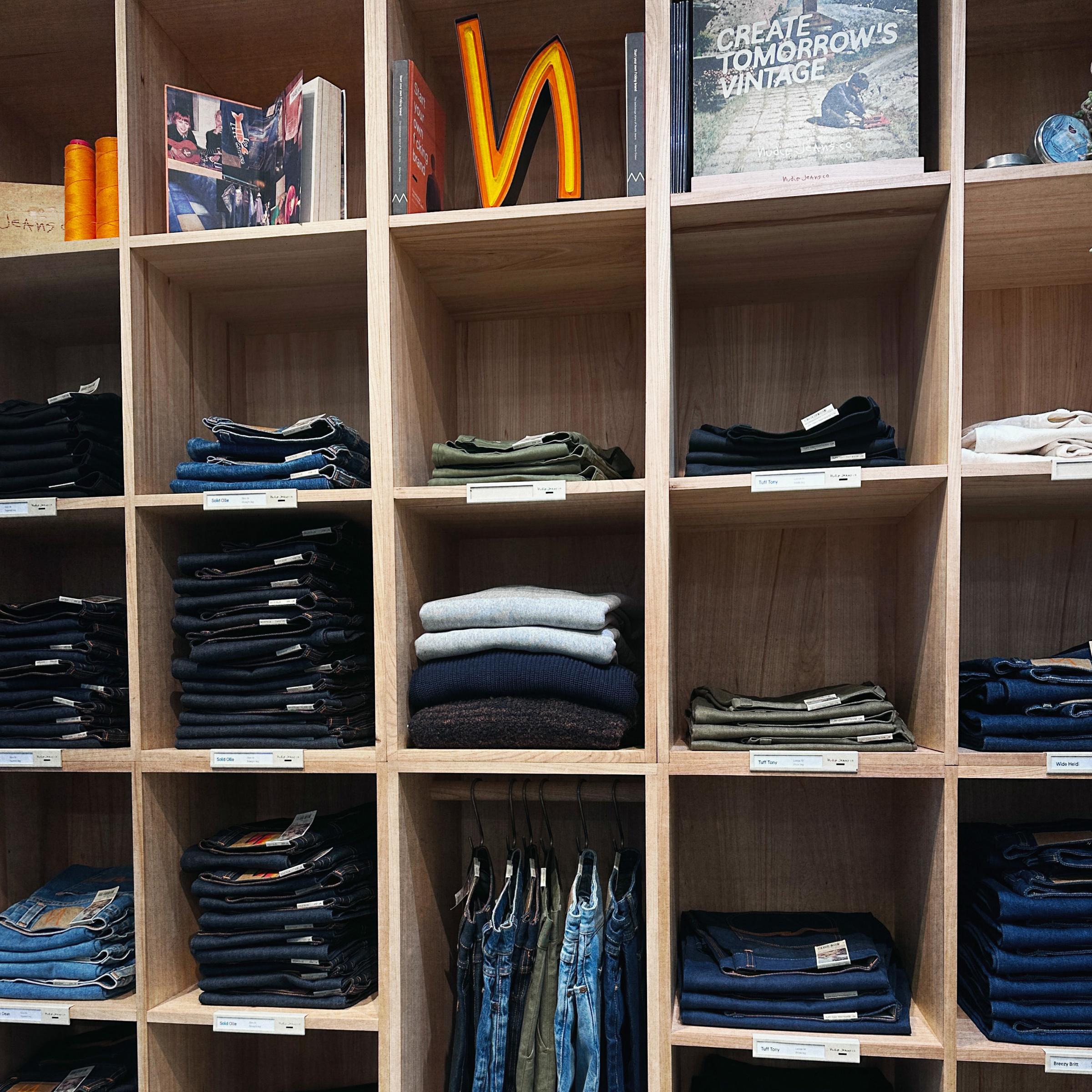 Neatly folded jeans and sweaters organized in wooden shelves with books and signs on top. Tags label each section.