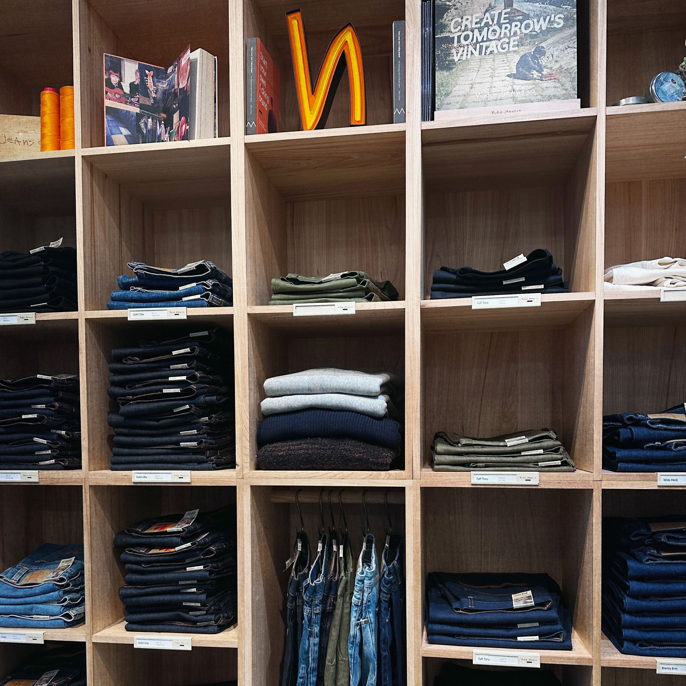 Neatly folded jeans and sweaters organized in wooden shelves with books and signs on top. Tags label each section.