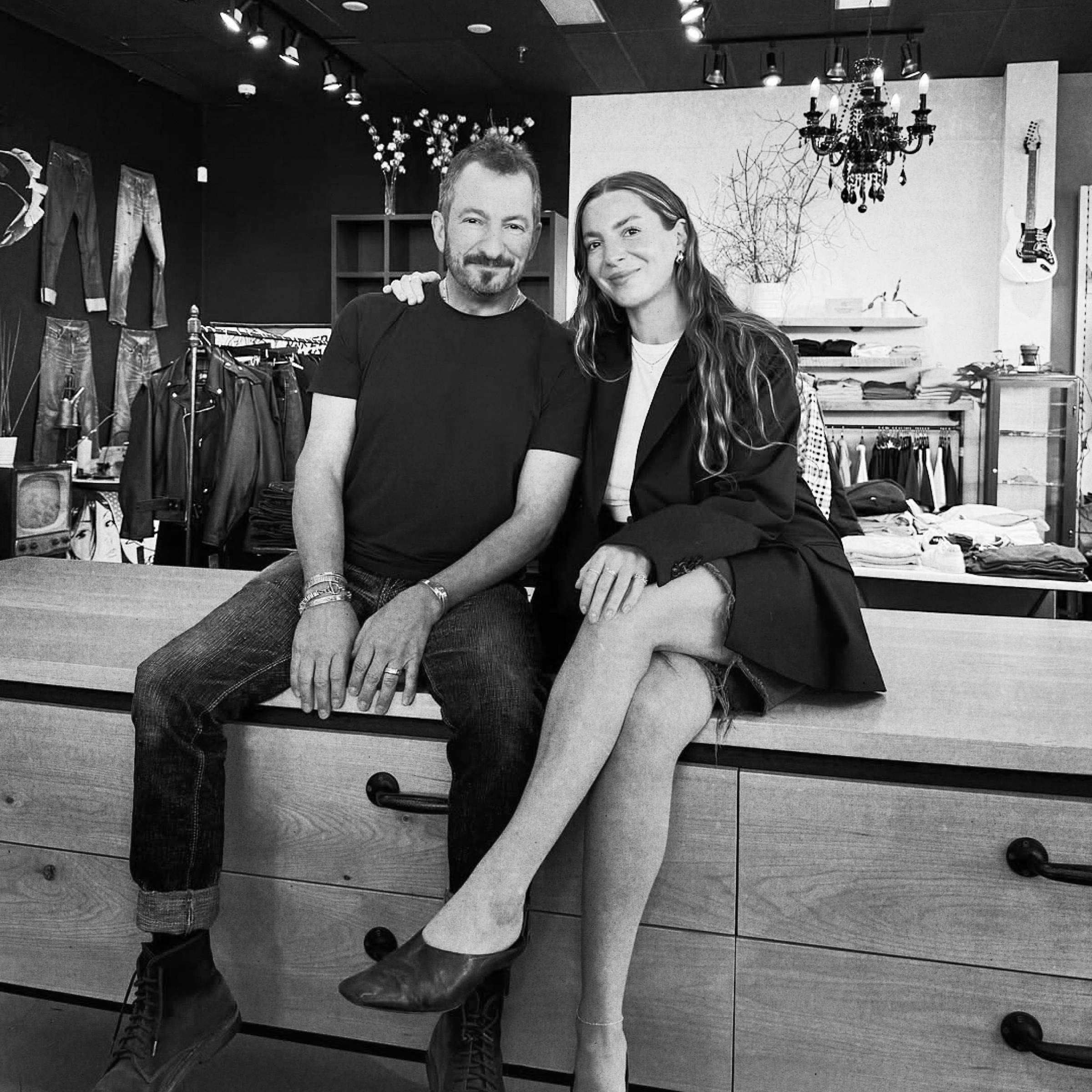 Aubrey and Jessie Margolis — the father–daughter duo behind Danali, sit on a wooden surface in a clothing store, smiling. The background displays clothes and decor on a black wall.