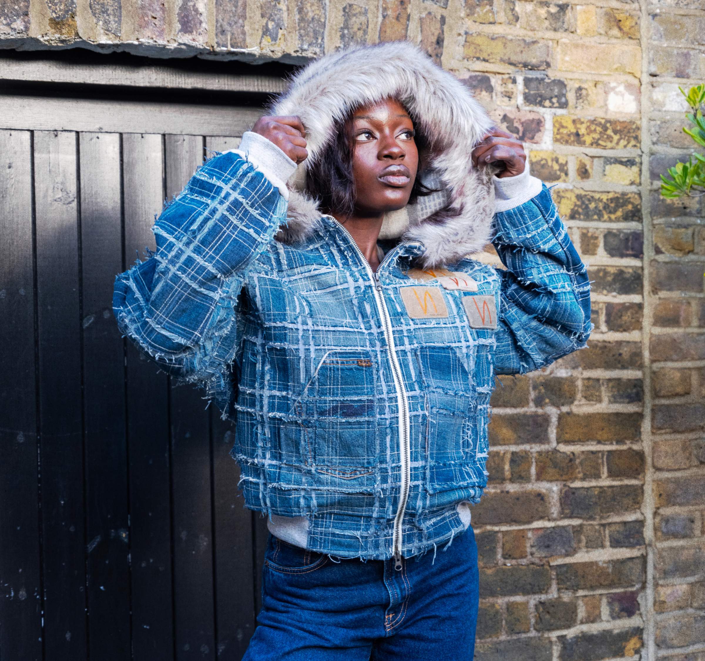 Designed bu Maco. Person wearing blue patchwork jacket with fur-lined hood, holding hood up against a brick wall and dark door.