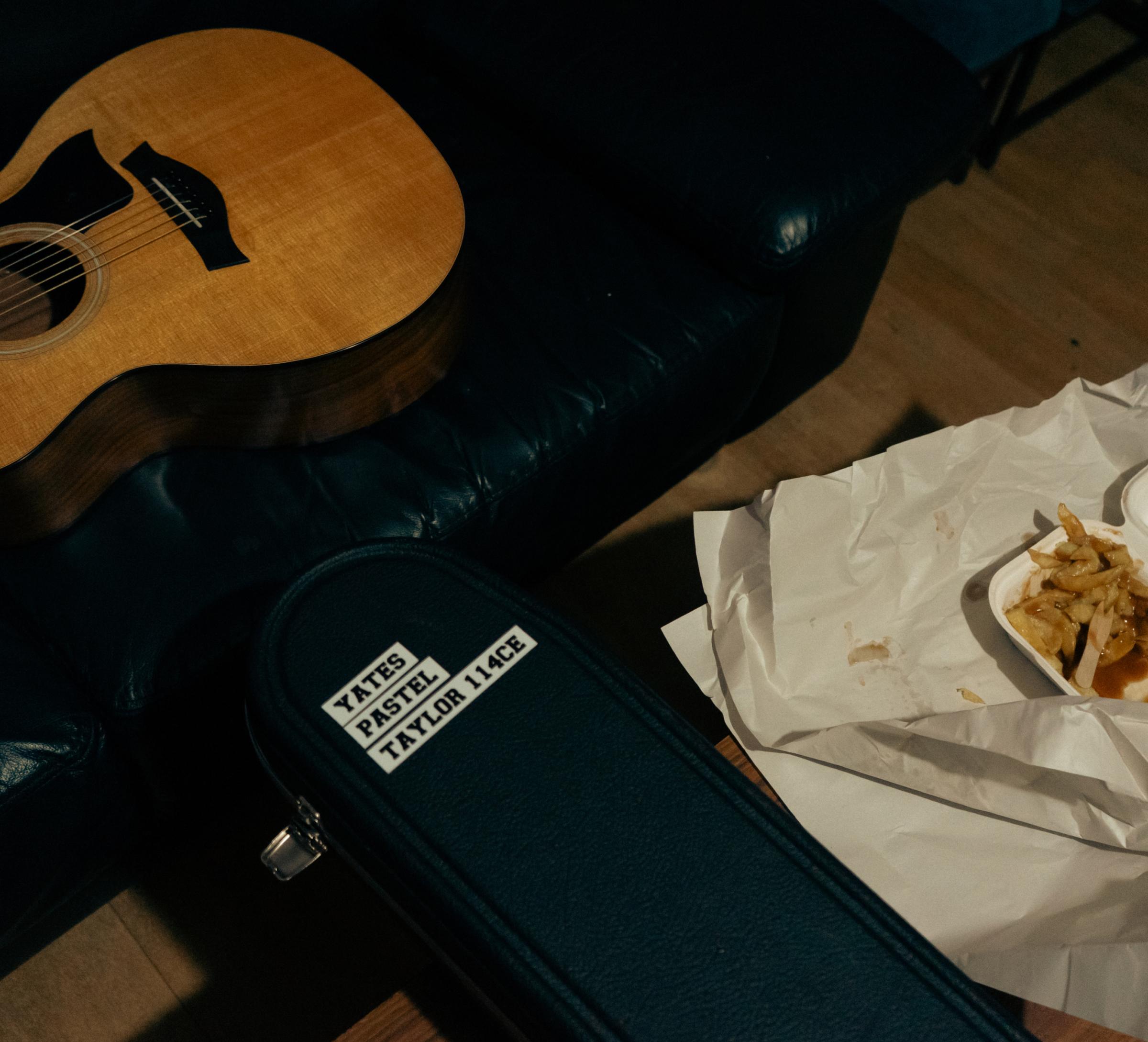 Acoustic guitar on a dark leather couch, a guitar case labeled "Yates Pastel Taylor 114CE", and takeout fries on crumpled paper.