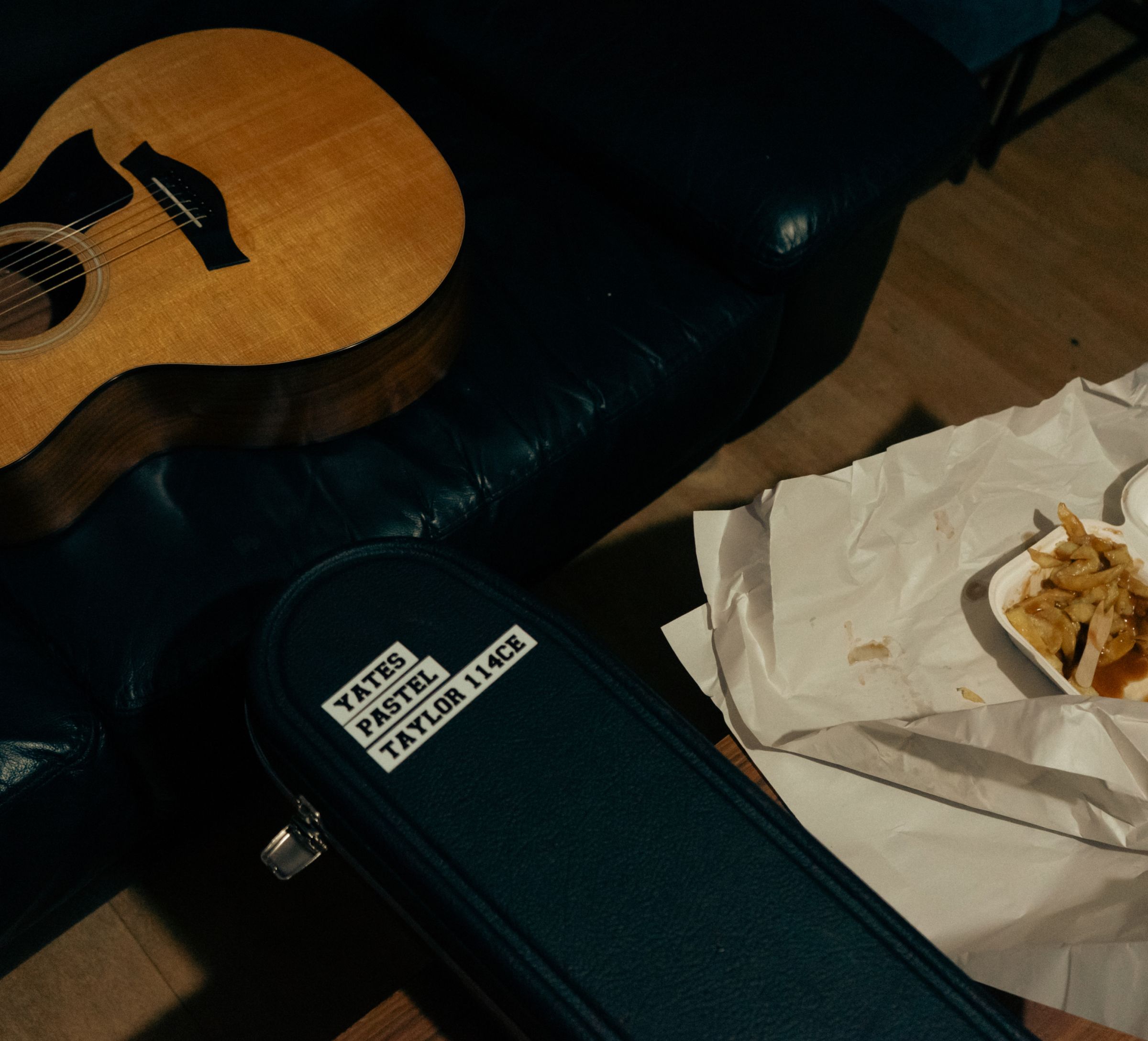 Acoustic guitar on a dark leather couch, a guitar case labeled "Yates Pastel Taylor 114CE", and takeout fries on crumpled paper.