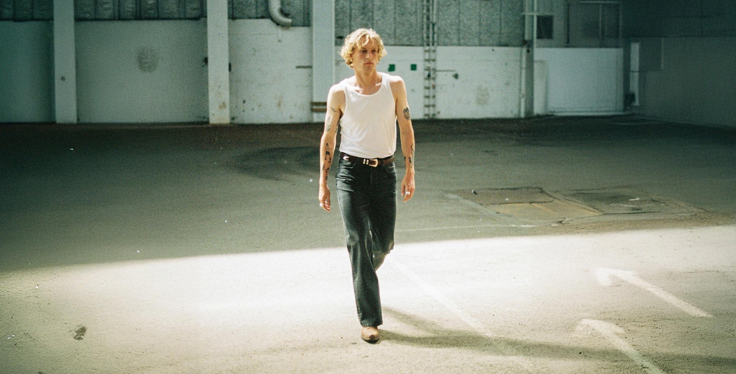 Person in a white tank top and black pants walking in a sunlit, empty industrial space.